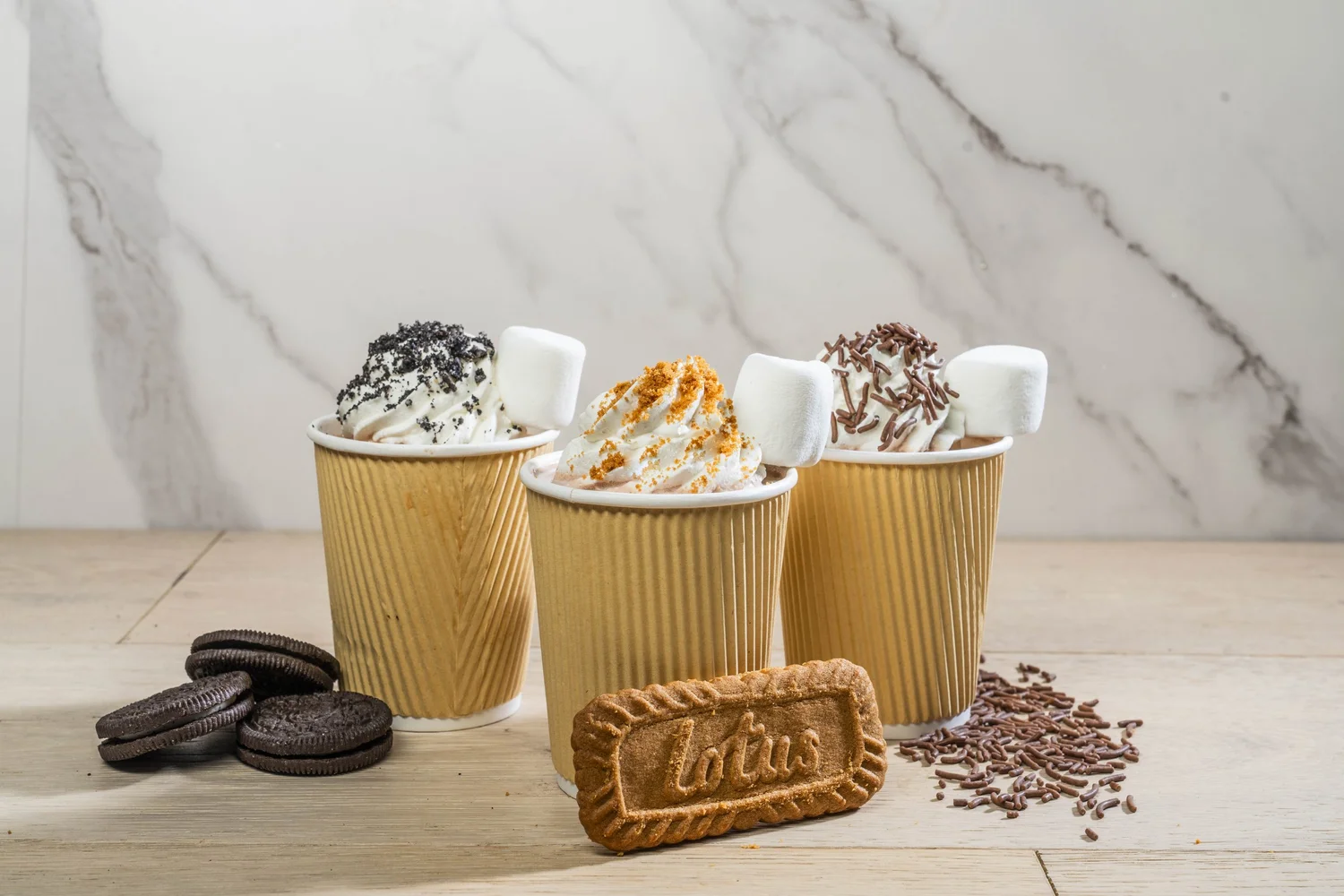 Three cups of hot chocolate in kraft sleeves with whipped cream, cookies and chocolate on marble