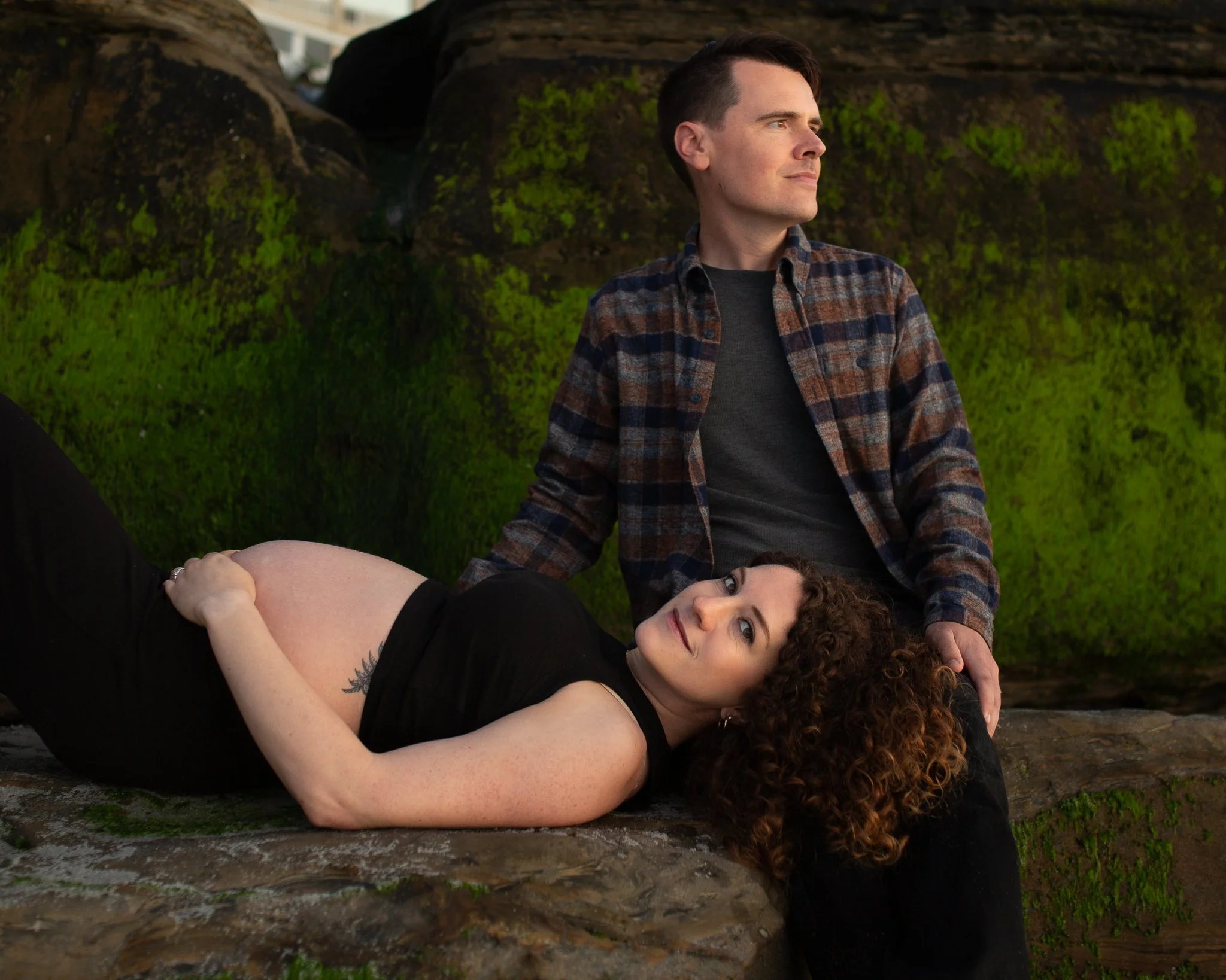A man and a woman pose outdoors near rocks and green moss. The man is sitting on top of the rocks, wearing a plaid shirt, and looking to the side. The woman with curly hair is lying on her side on the rocks, looking up at the camera, wearing a black top.