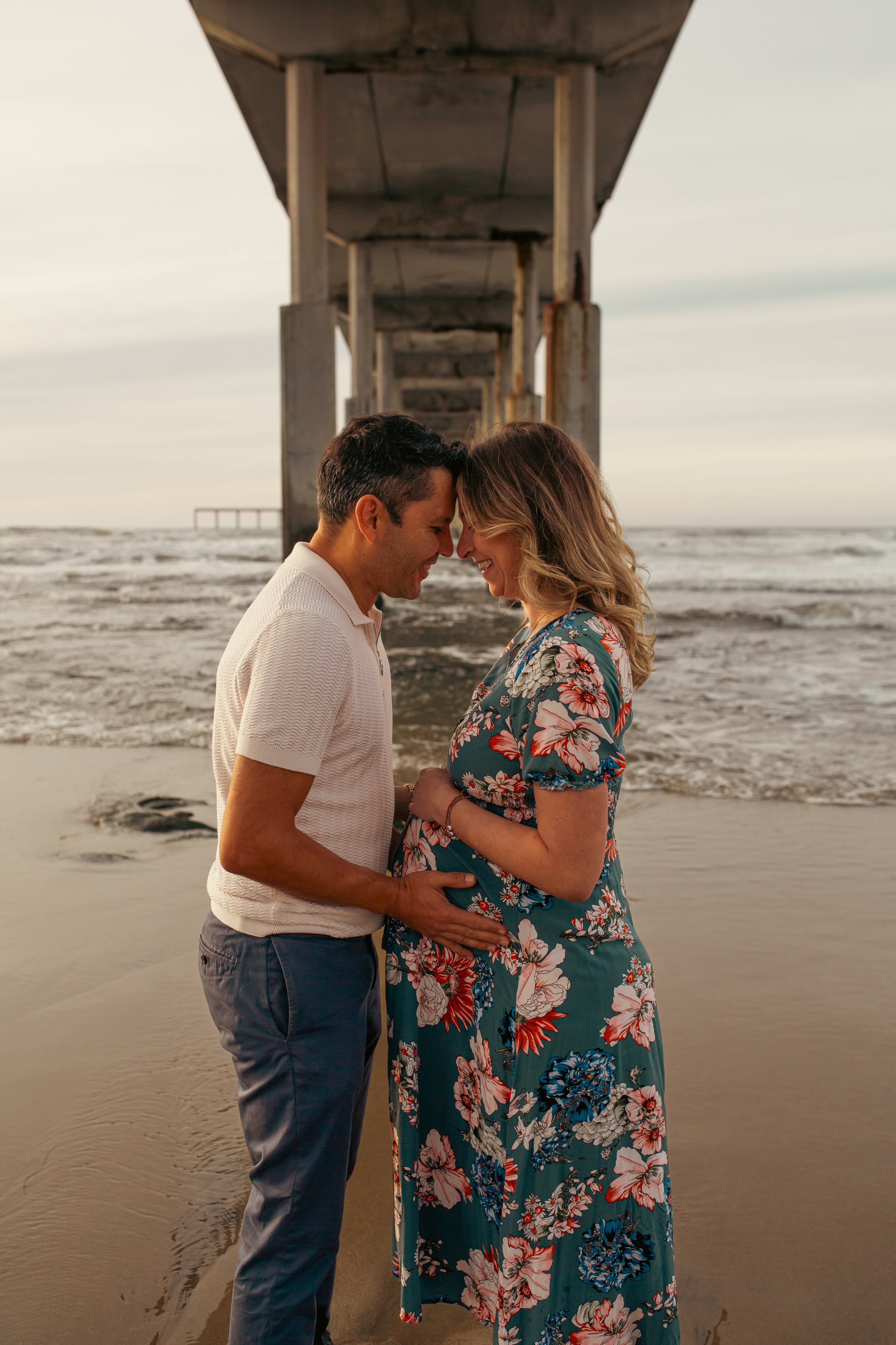 A couple stands close with foreheads touching on the beach under a pier at sunset, smiling at each other, with ocean waves in the background.