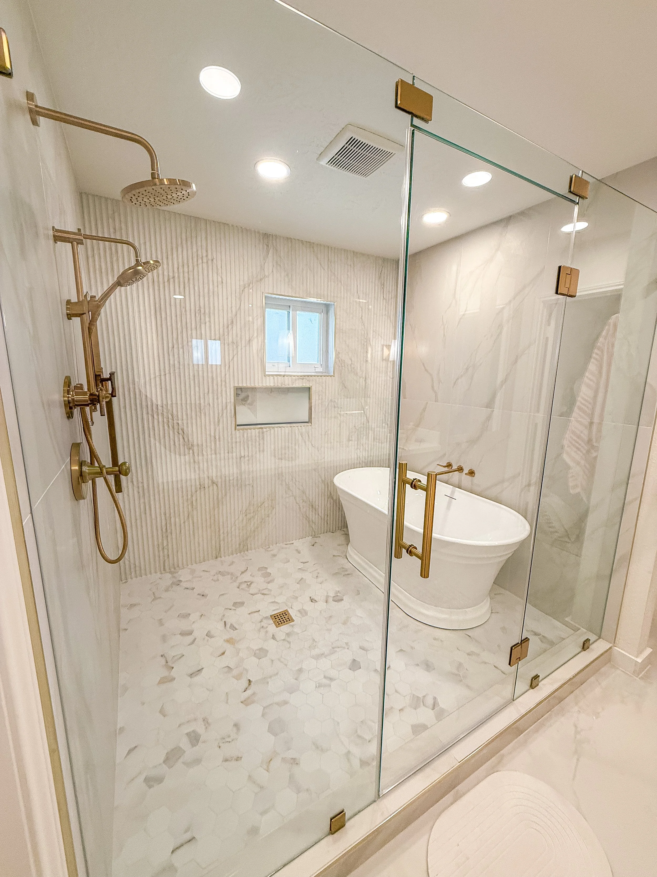 Modern bathroom with a glass shower door, gold fixtures, marble walls, two showerheads, a niche with a window, a freestanding bathtub, and a hexagonal tile floor.