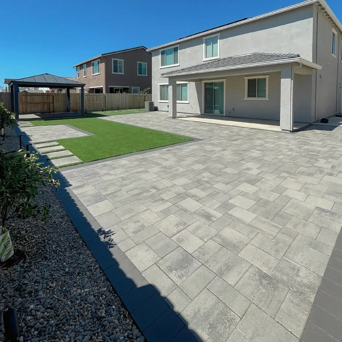 A backyard with a large paved patio area, a small patch of artificial grass with stepping stones, a wooden fence, a gazebo, and a two-story house with a covered patio overlooking the yard.