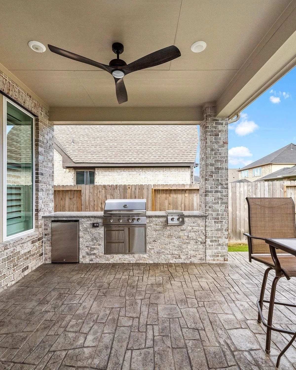 Covered outdoor patio with brick walls, ceiling fan, and built-in stainless steel grill and mini-fridge, overlooking a backyard with wooden fence and neighboring houses.