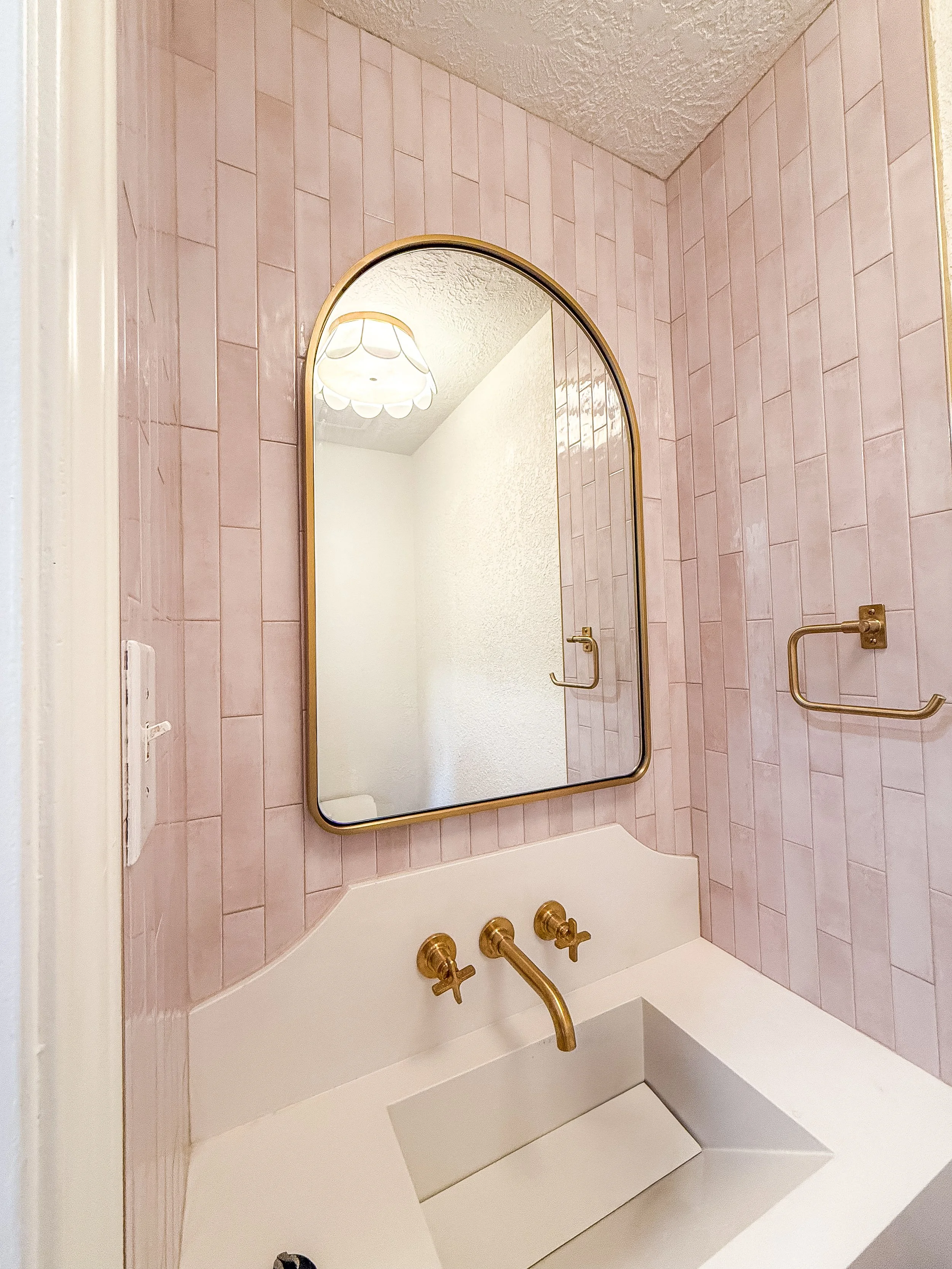 A bathroom with pink tiled walls, a large arched mirror with a gold frame, brass towel bars, and a white bathtub with brass fixtures and a grab bar.