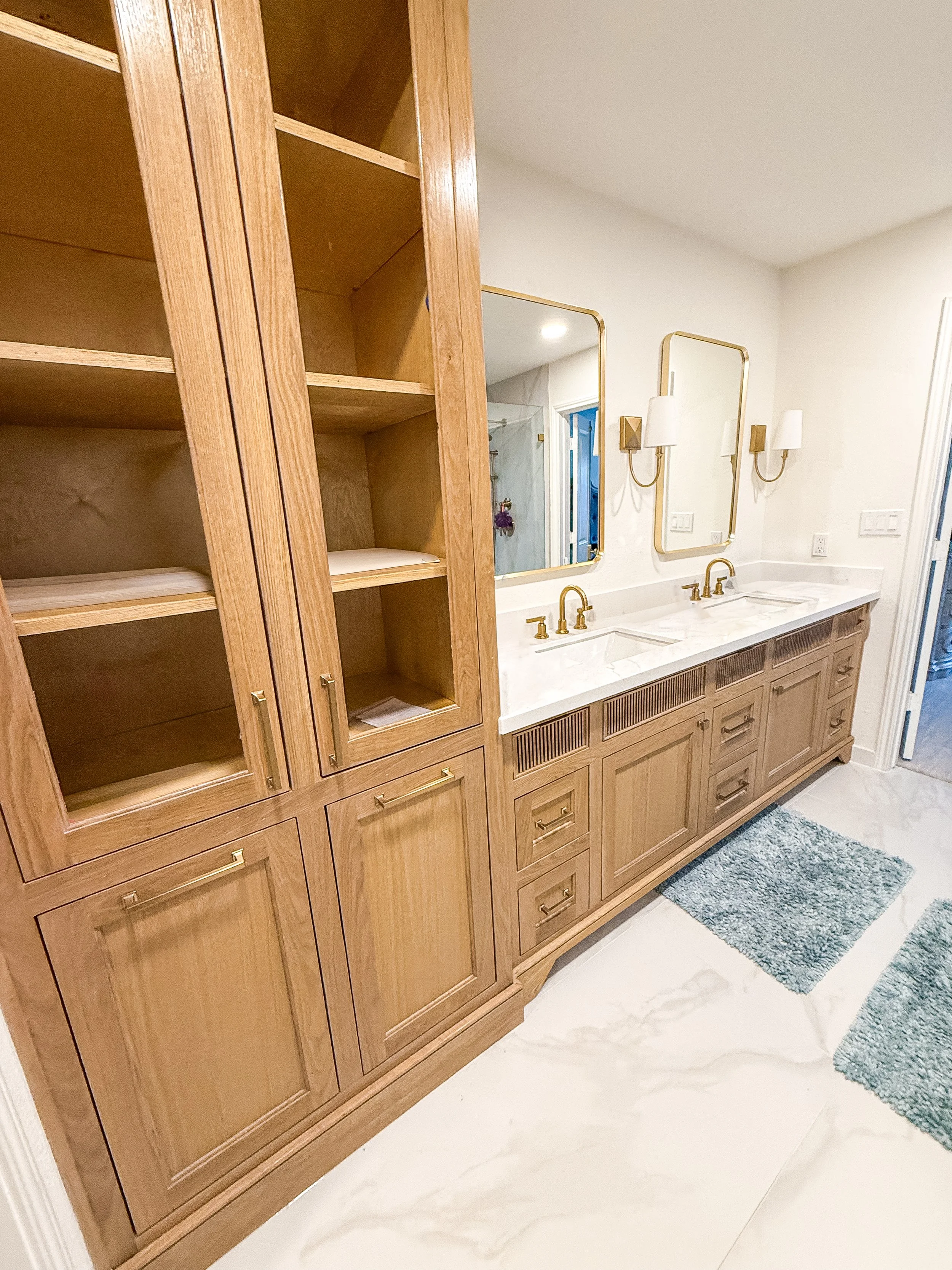 Bathroom with wooden cabinets, marble countertop, two mirrors, and wall-mounted lights, with two blue rugs on white marble floor.