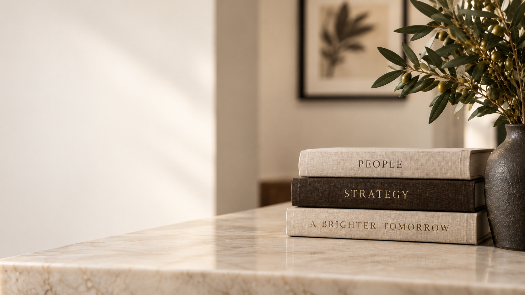 A stack of three hardcover books titled "People," "Strategy," and "A Brighter Tomorrow" on a marble tabletop. To the right, a dark-gray textured vase holding green leafy branches. In the background, a framed black-and-white botanical print hangs on a light-colored wall.