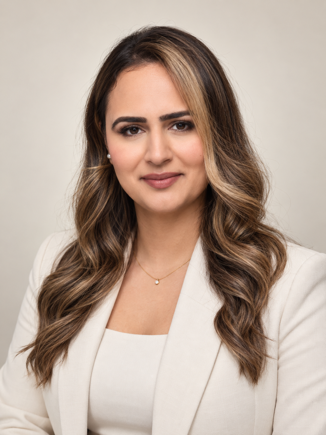 Portrait of a woman with long, wavy brown hair and light skin, wearing a white blazer and a delicate necklace with a small pearl pendant, against a plain light background.
