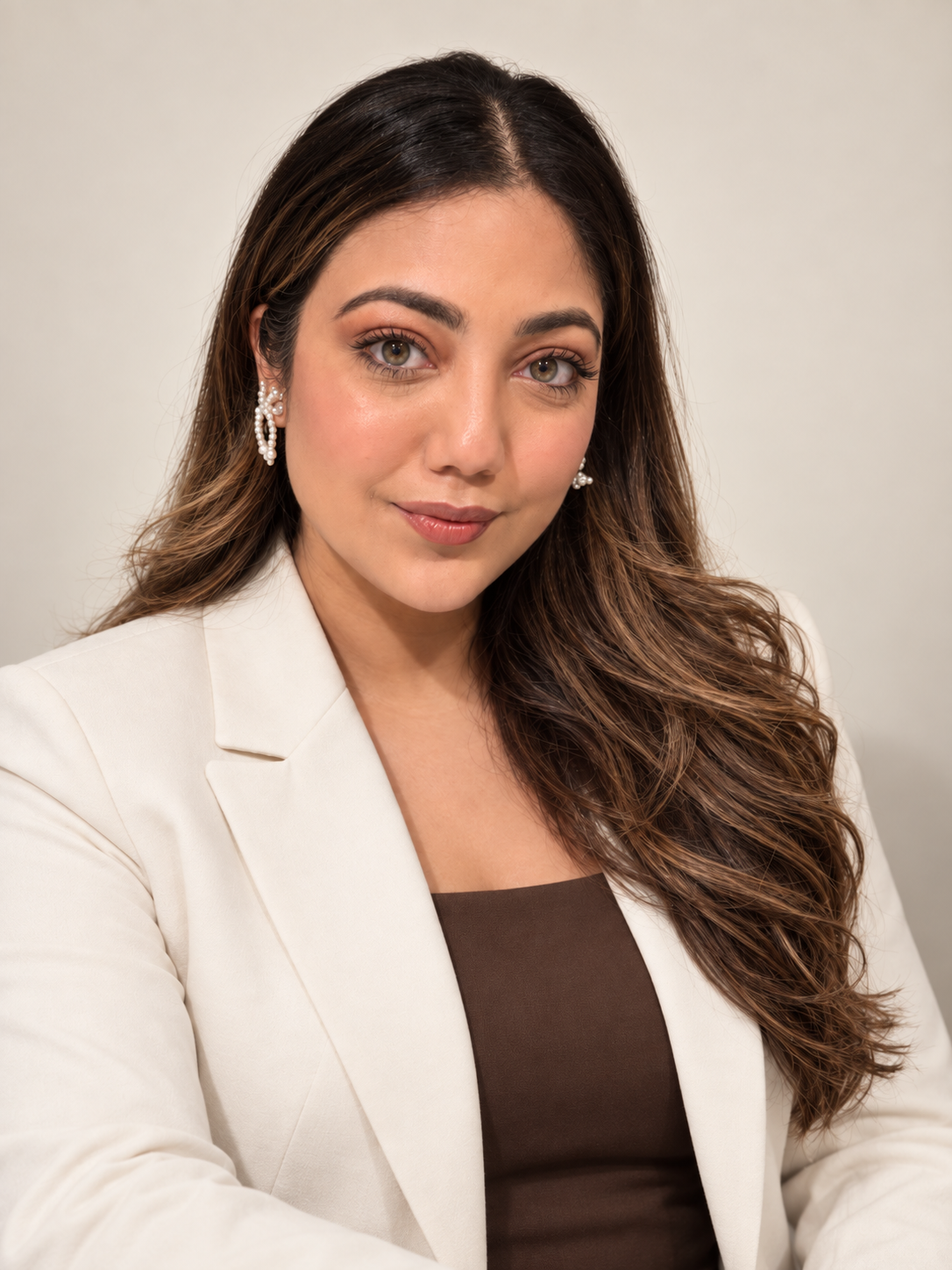 A woman with long, wavy brown hair, wearing pearl earrings and a white blazer over a brown top, looking at the camera with a slight smile against a plain light background.