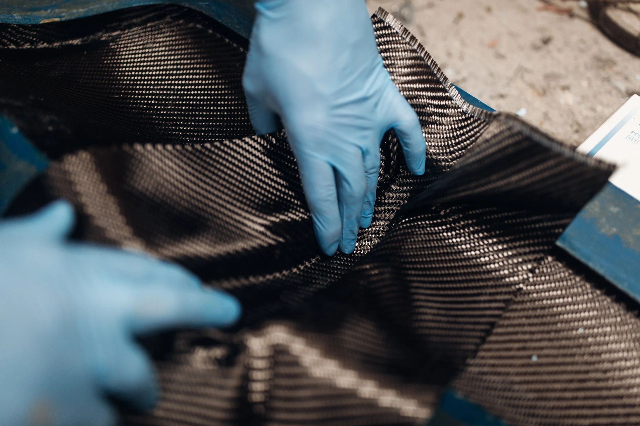 Close-up of a hand wearing a blue glove touching a black and white patterned fabric, possibly a carbon fiber material, with a blurred background.