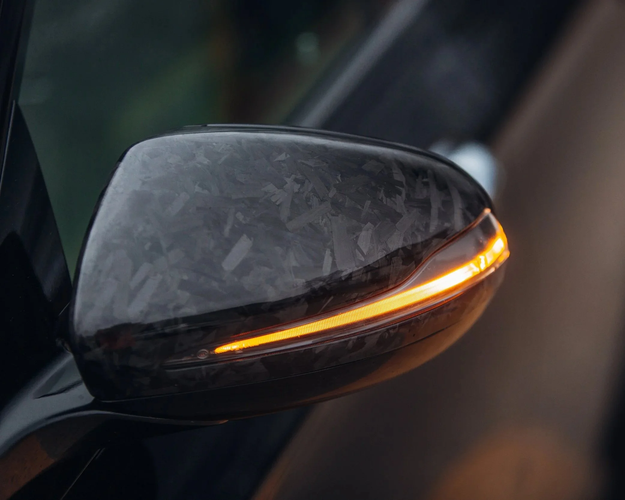 Close-up of a black vehicle's front left side, showing a part of the headlight with an orange indicator light on in a dimly lit setting.