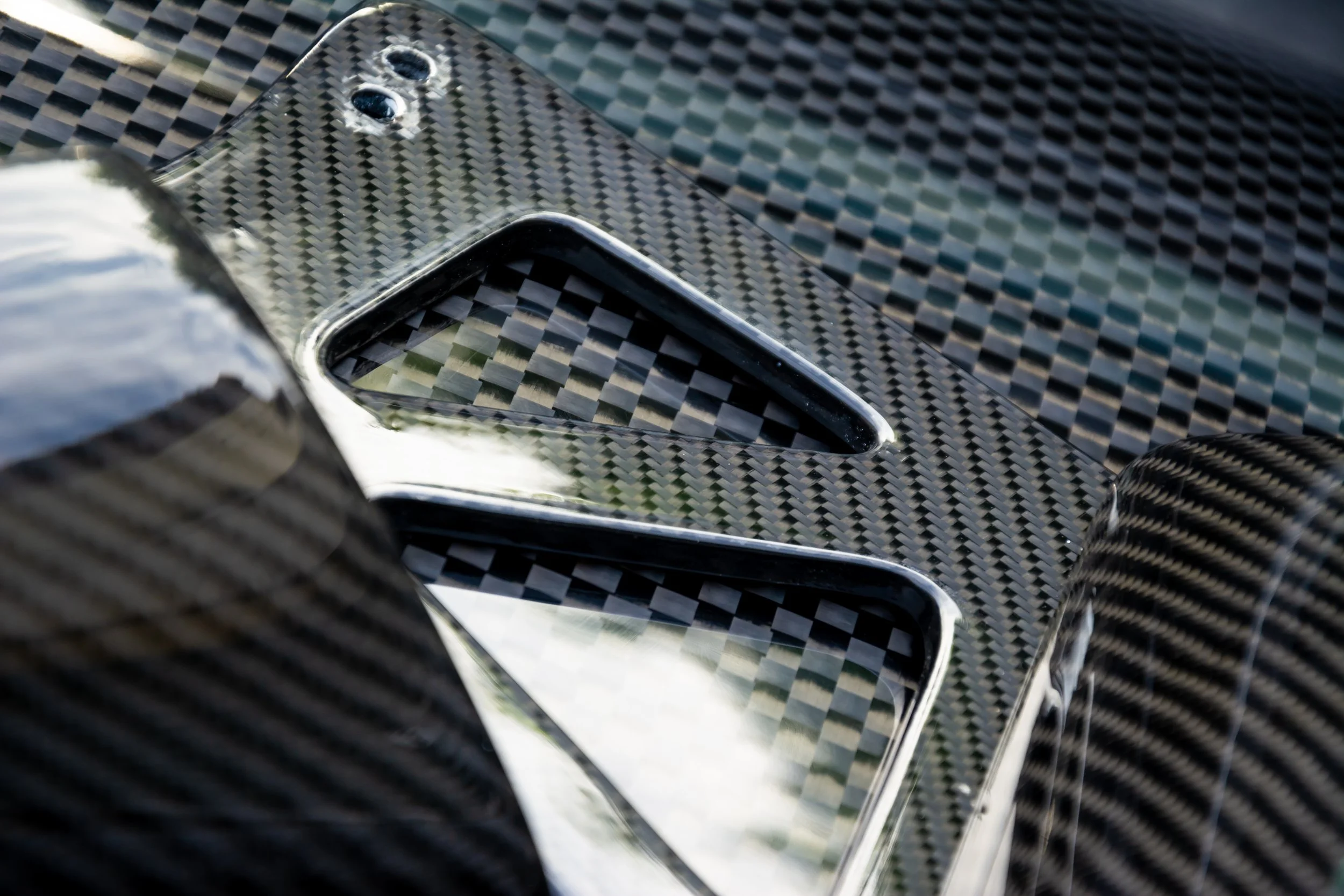 Close-up of a reflective carbon fiber surface with an opening, possibly the back of a high-performance car or device, showing a textured pattern and shiny finish.