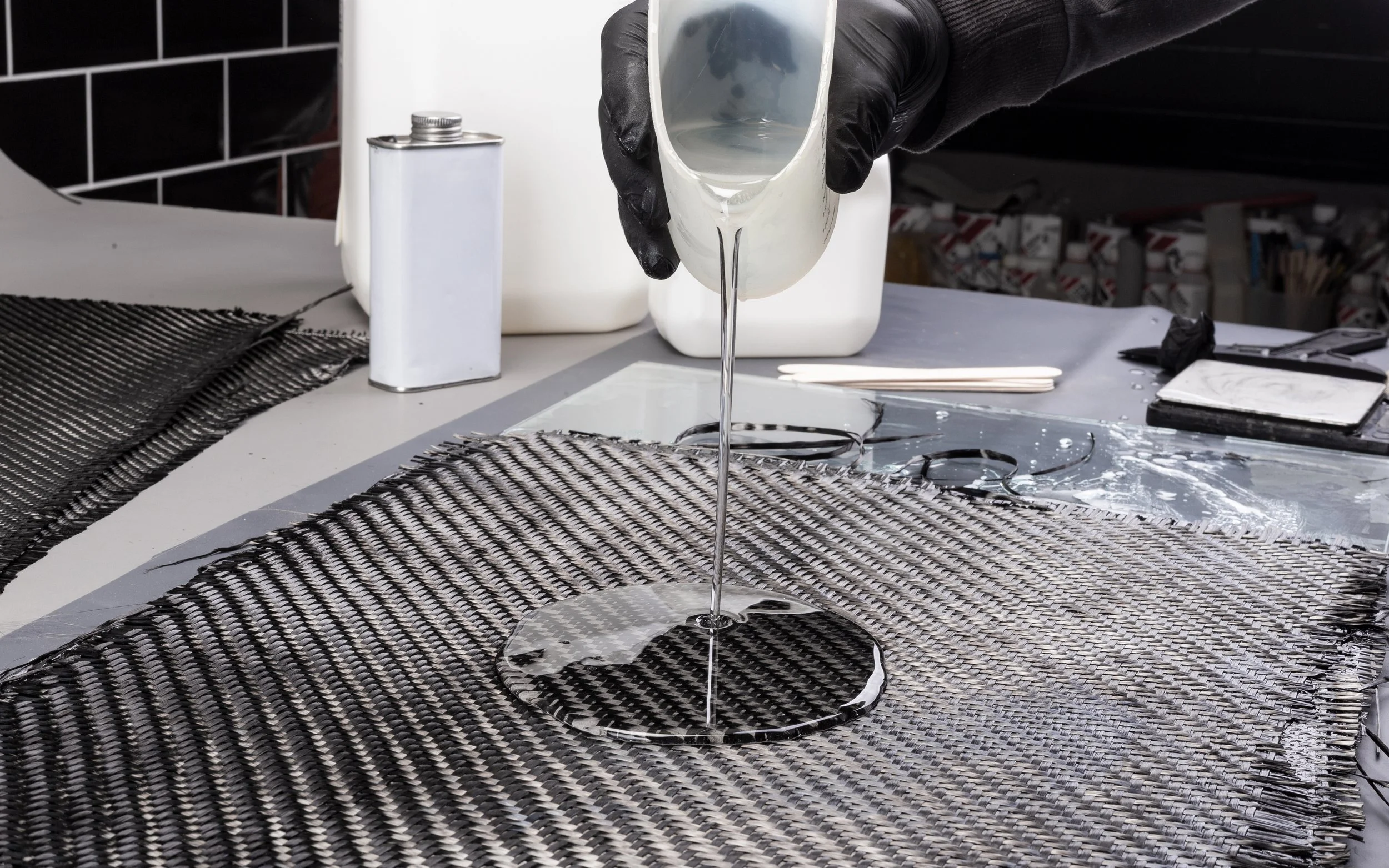 A person's hand wearing a black glove pouring a clear liquid from a small container onto a black and white woven mat on a table in a workshop or manufacturing setting.