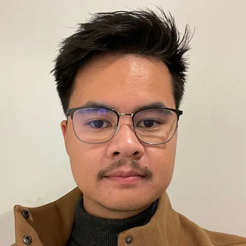 Portrett av en mann med briller, kort mørkt hår, og faux-mustasje, kledd i en brun jakke og svart genser, mot en hvit bakgrunn.