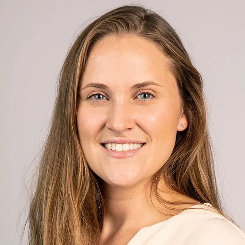 Et nærbilde av en smilende kvinne med langt brunt hår og lyse øyne, iført en lysfarget topp, mot en enkel grå bakgrunn.