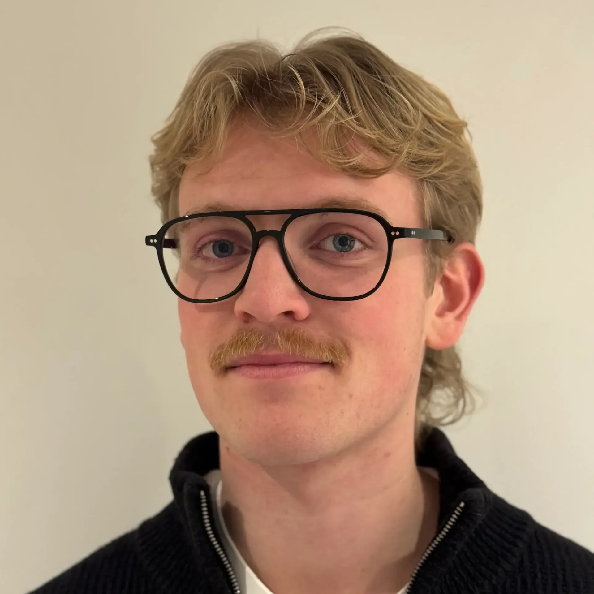 Nærbilde av ansiktet til en mann med blond, krøllete hår, briller og skjegg, iført en svart hettegenser med glidelås.