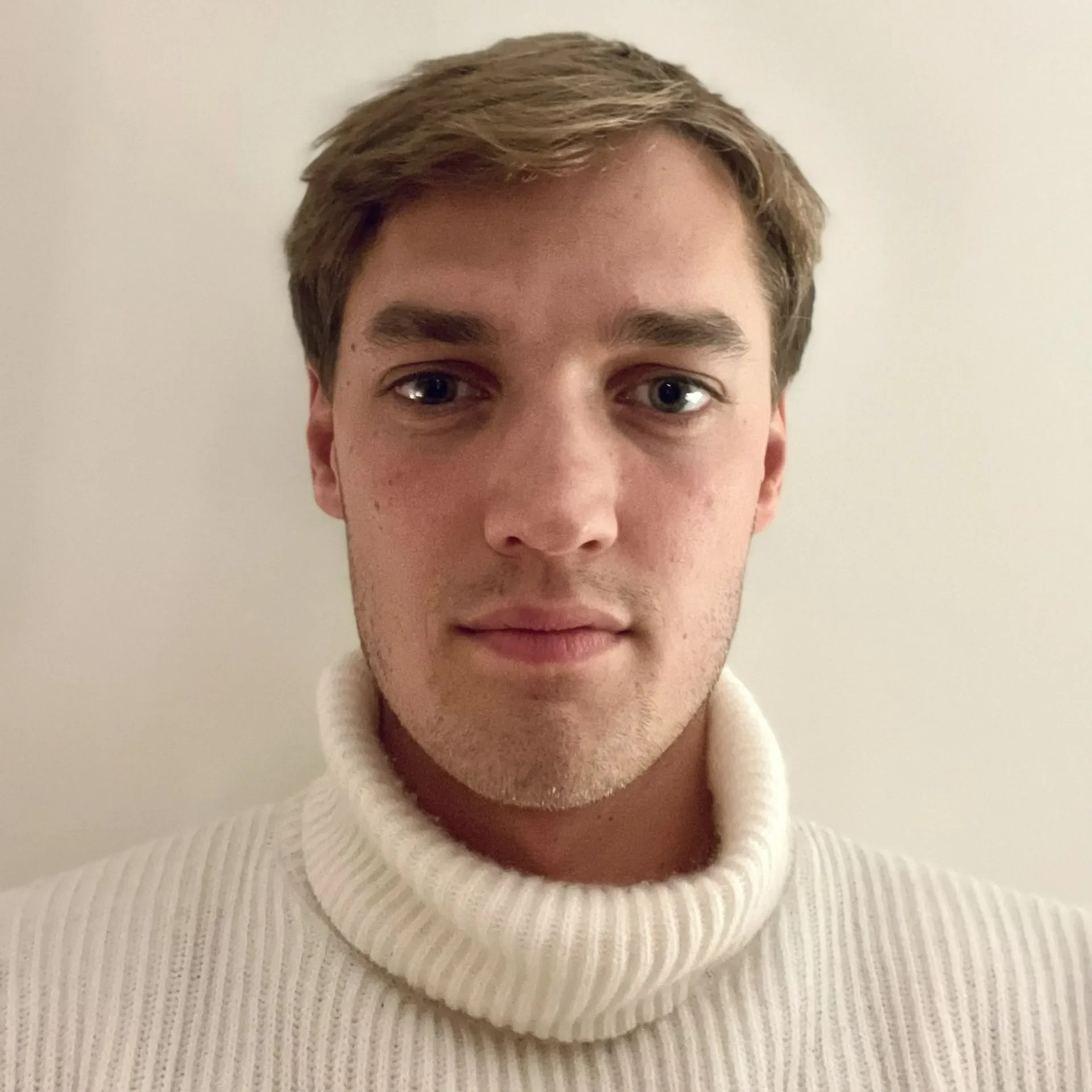 Et portrett av en ung mann med lyst, rutete hår, smilende, iført en hvit strikket genser, foran en nøytral lys bakgrunn.