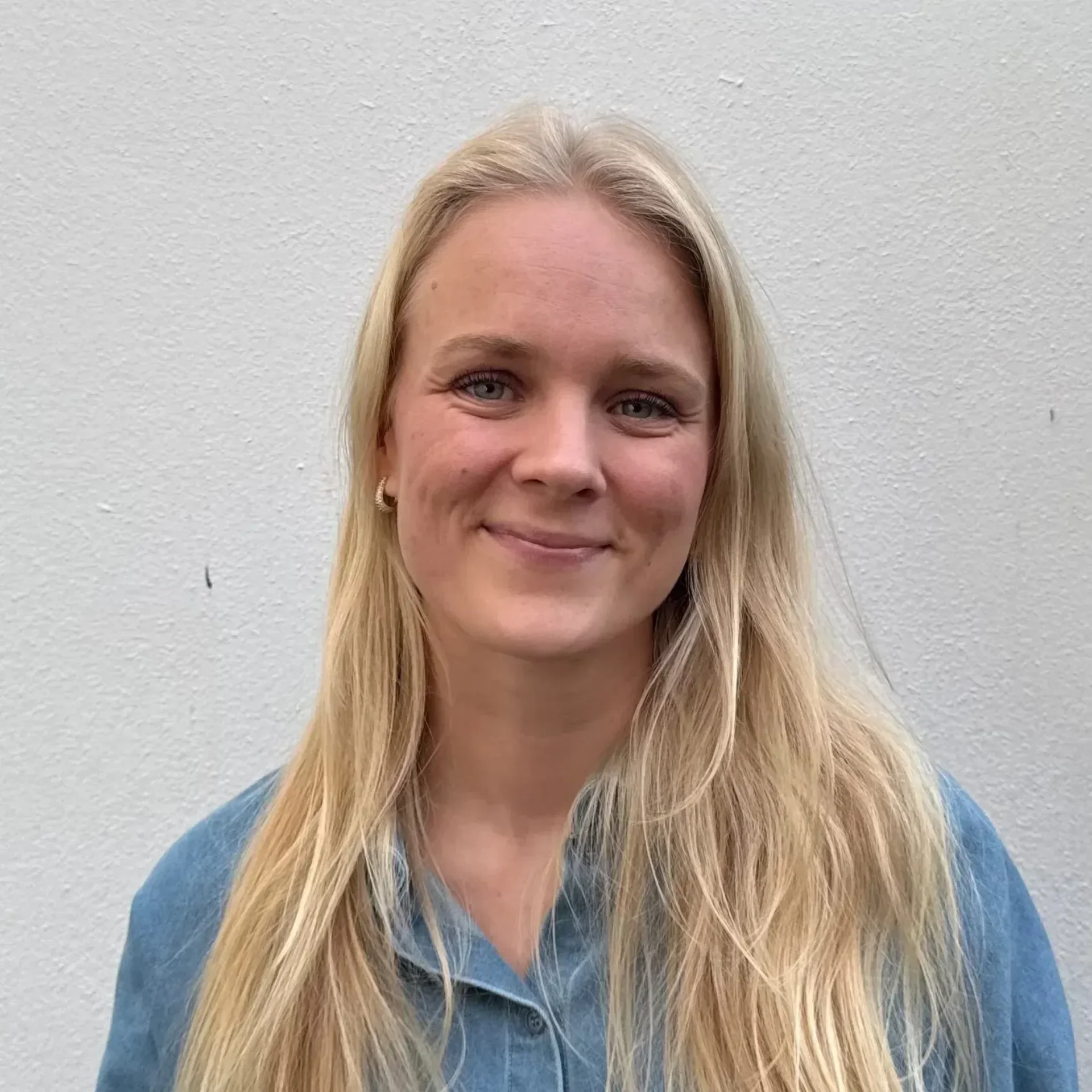 Nærbilde av en smilende kvinne med langt blonde hår, iført blå genser, foran en hvit vegg.