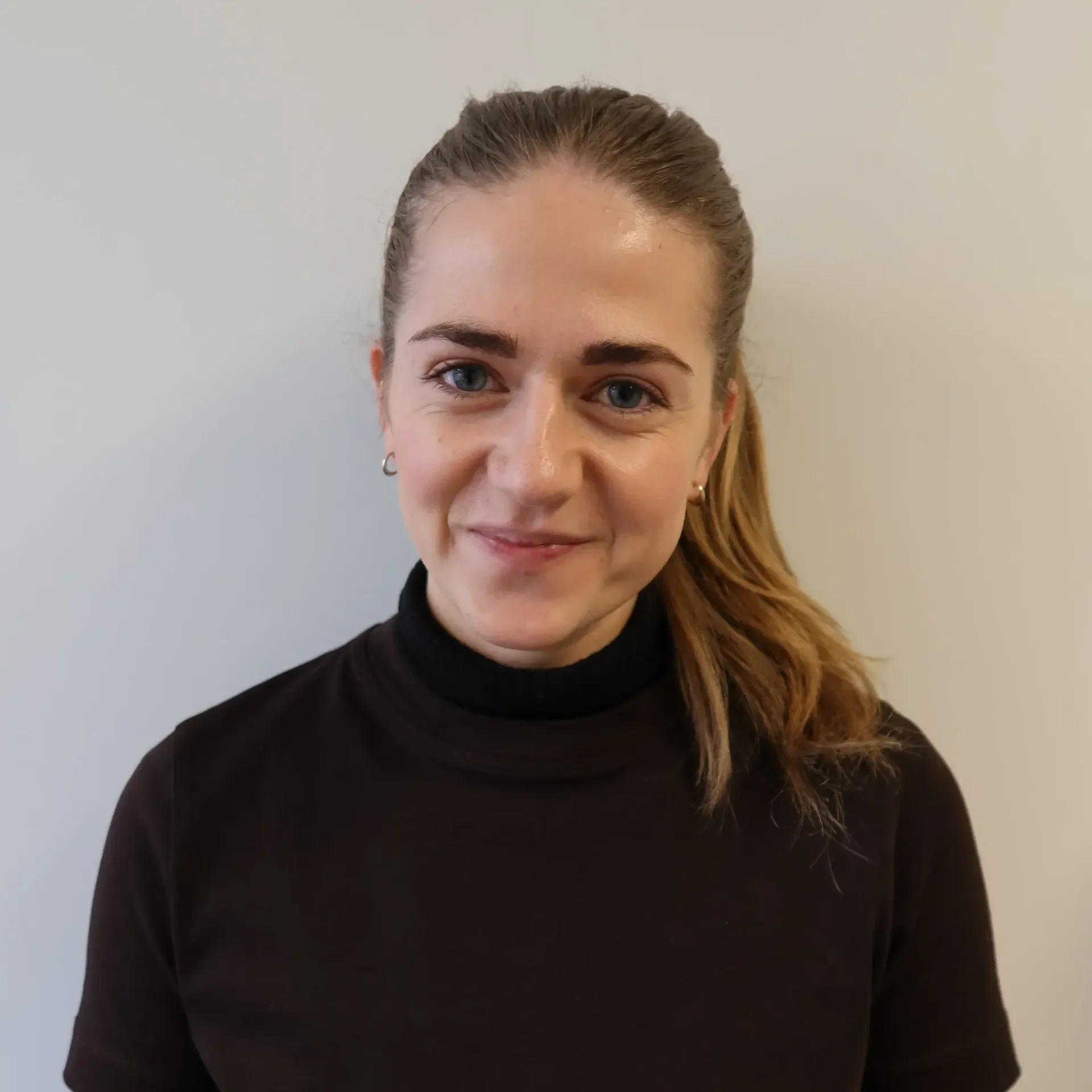 Nærbilde av en smilende kvinne med langt, blond hår, iført svart genser, foran en lys vegg.