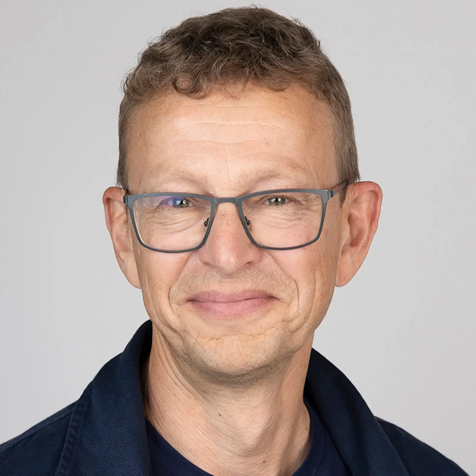Nærbilde av en mann med briller og kort, krøllete hår som smiler, mot en grå bakgrunn.