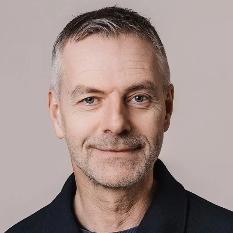Portrettbilde av en mann med kort grått hår, lys hudfarge og et halvt smil, iført en sort jakke, mot en lys bakgrunn.