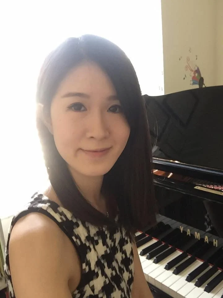 Young woman with dark hair smiling in front of a Yamaha grand piano, wearing a patterned sleeveless top.