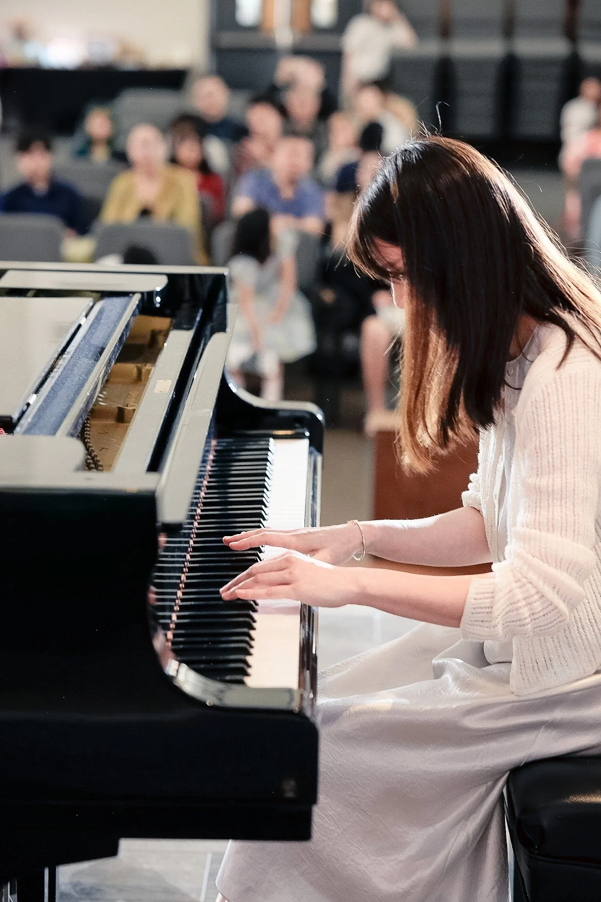 A woman with shoulder-length dark hair playing the piano in front of an audience in a dimly lit room.