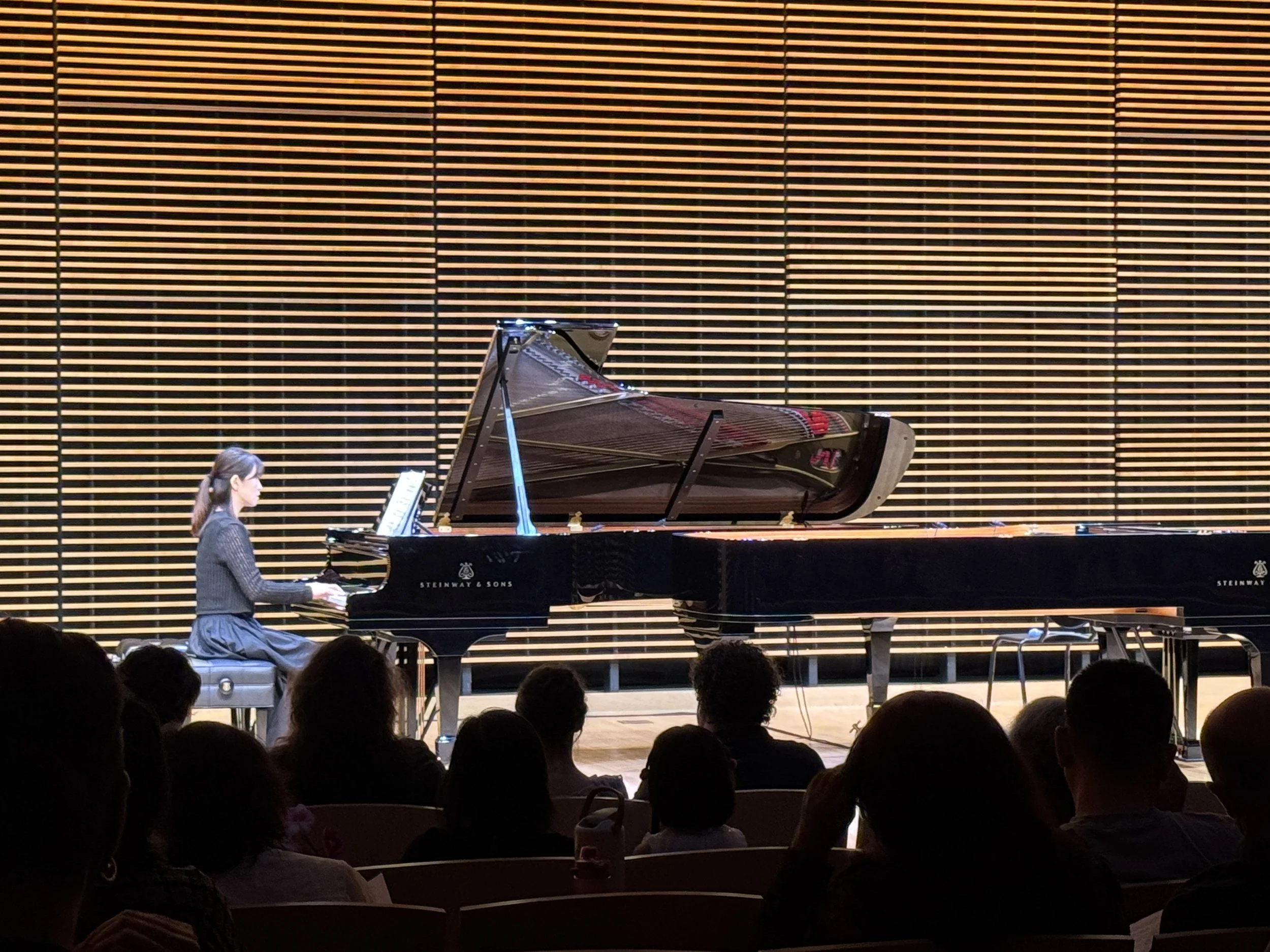 A woman playing a grand piano on stage in front of an audience, with a large wooden wall in the background.