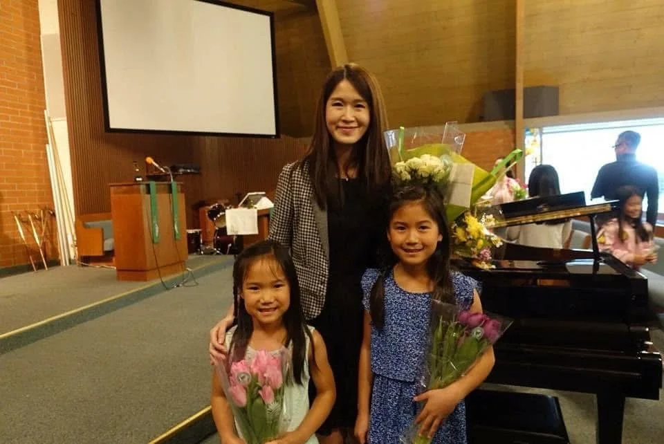 A woman and two young girls smiling inside a room with a piano, holding bouquets of flowers.