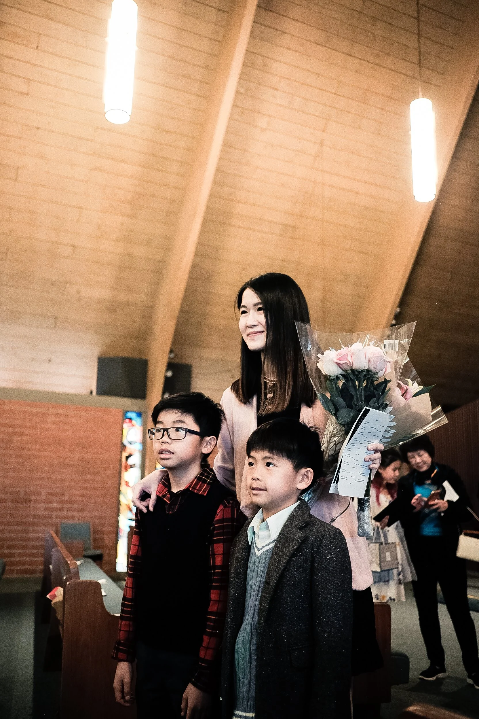 A woman with long dark hair smiling and holding a bouquet of pink and white roses is standing behind three children, all of whom are looking forward. The woman is wearing a light pink outfit, and the children are dressed in various jackets and sweaters. They are inside a building with wooden ceiling and brick wall, with other people in the background.