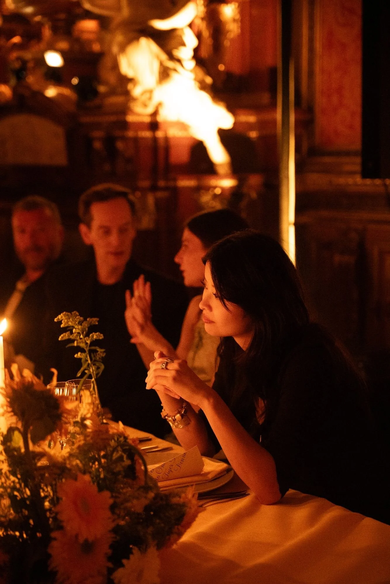 People dining in a dimly lit restaurant with a warm ambiance, decorated with flowers and lit by candles, a woman with black hair and bracelets is in the foreground, engaging in conversation while others are seated around the table.