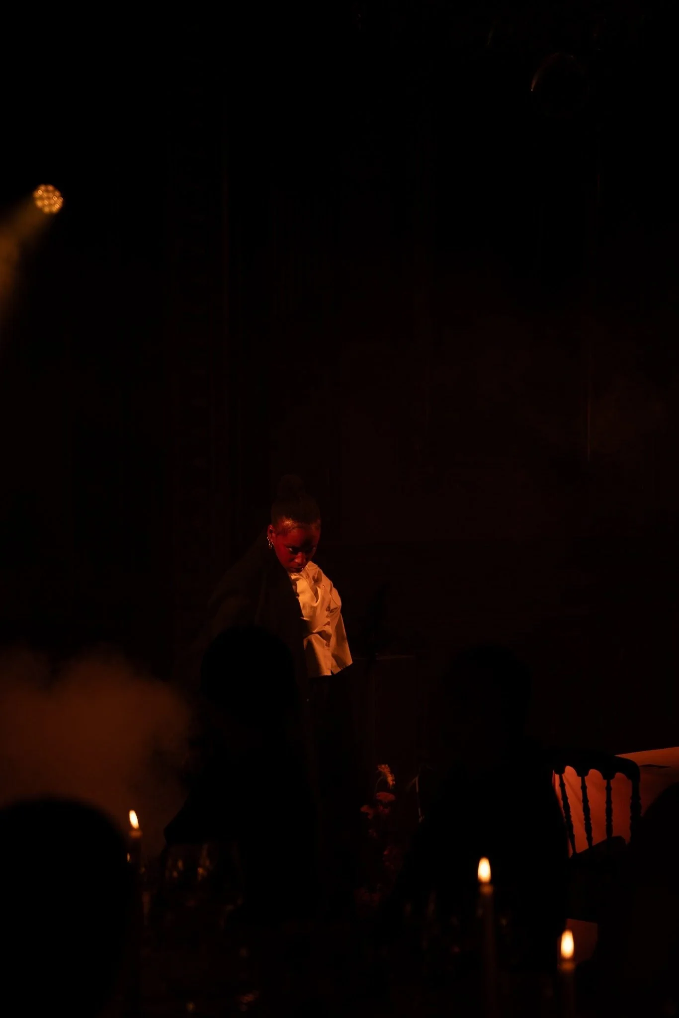 A woman wearing glasses and a black blazer is on stage, illuminated by warm stage lighting, with candles visible in the foreground and audience members in silhouette.