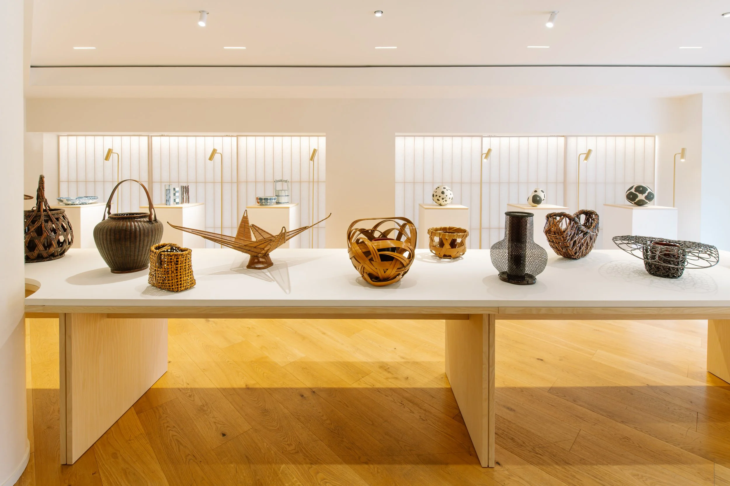 Display of various woven baskets and ceramics on a white table in a bright gallery space with wooden floors.