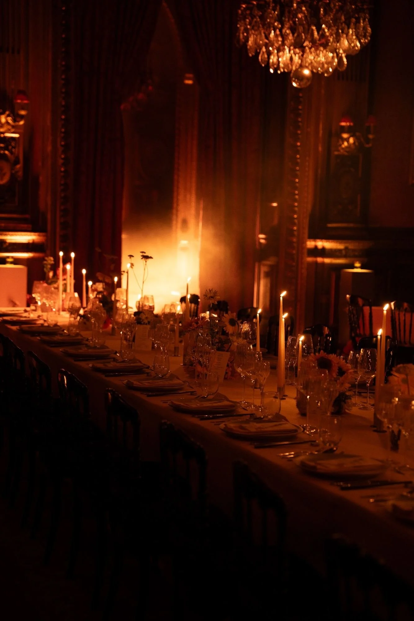 An elegant dining room with a long table set with glassware, plates, and candles. The room features ornate dark wooden paneling, a large chandelier, and dim warm lighting creating a sophisticated ambiance.