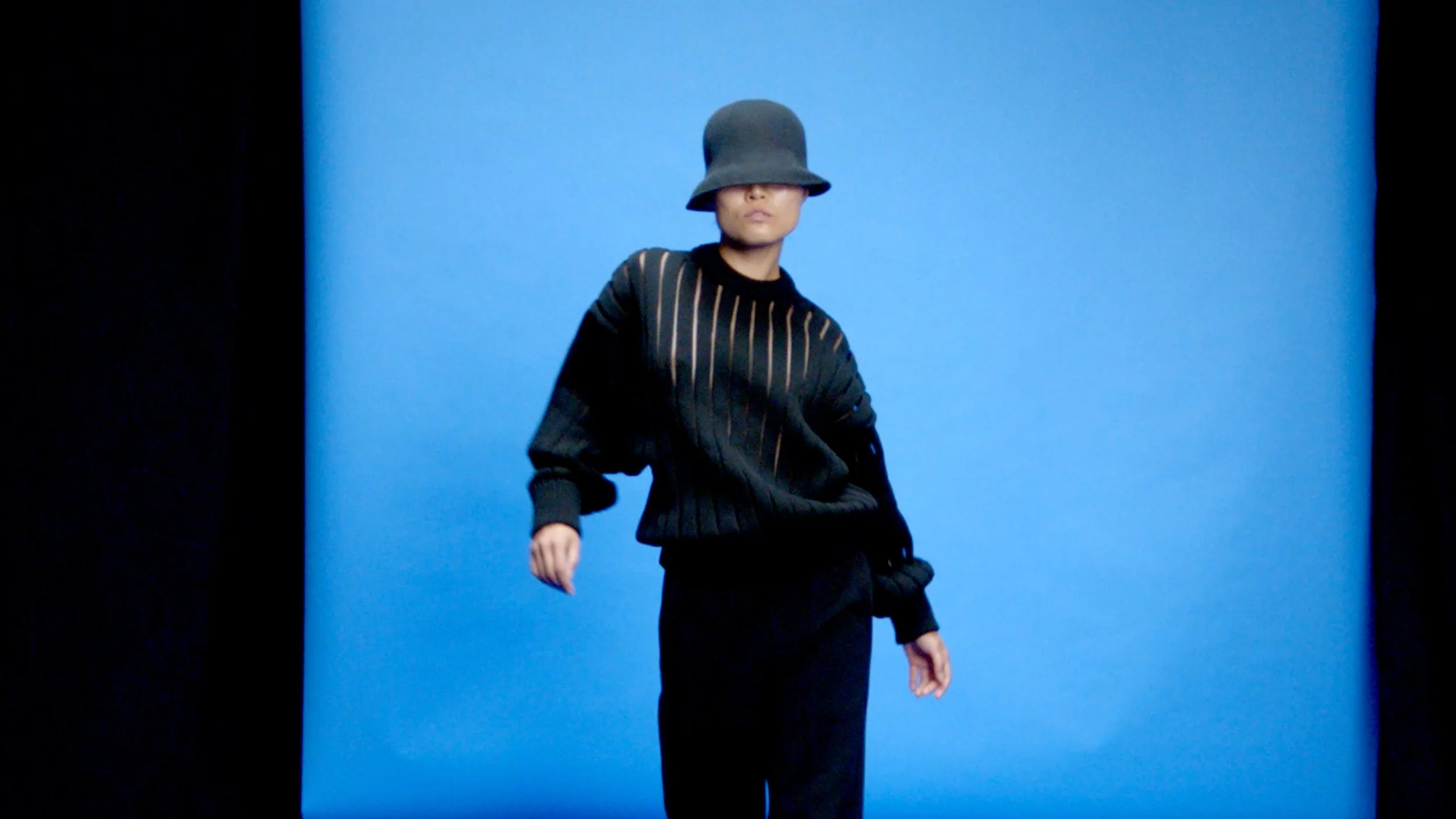 A person wearing a black hat and a black shirt with gold vertical stripes, standing against a blue background.