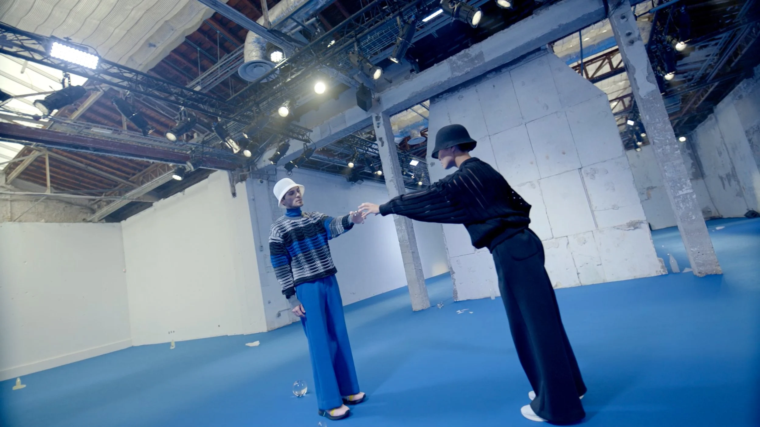 Two women in stylish clothing and hats practicing a dance move in an industrial-style studio with exposed ceilings and walls.