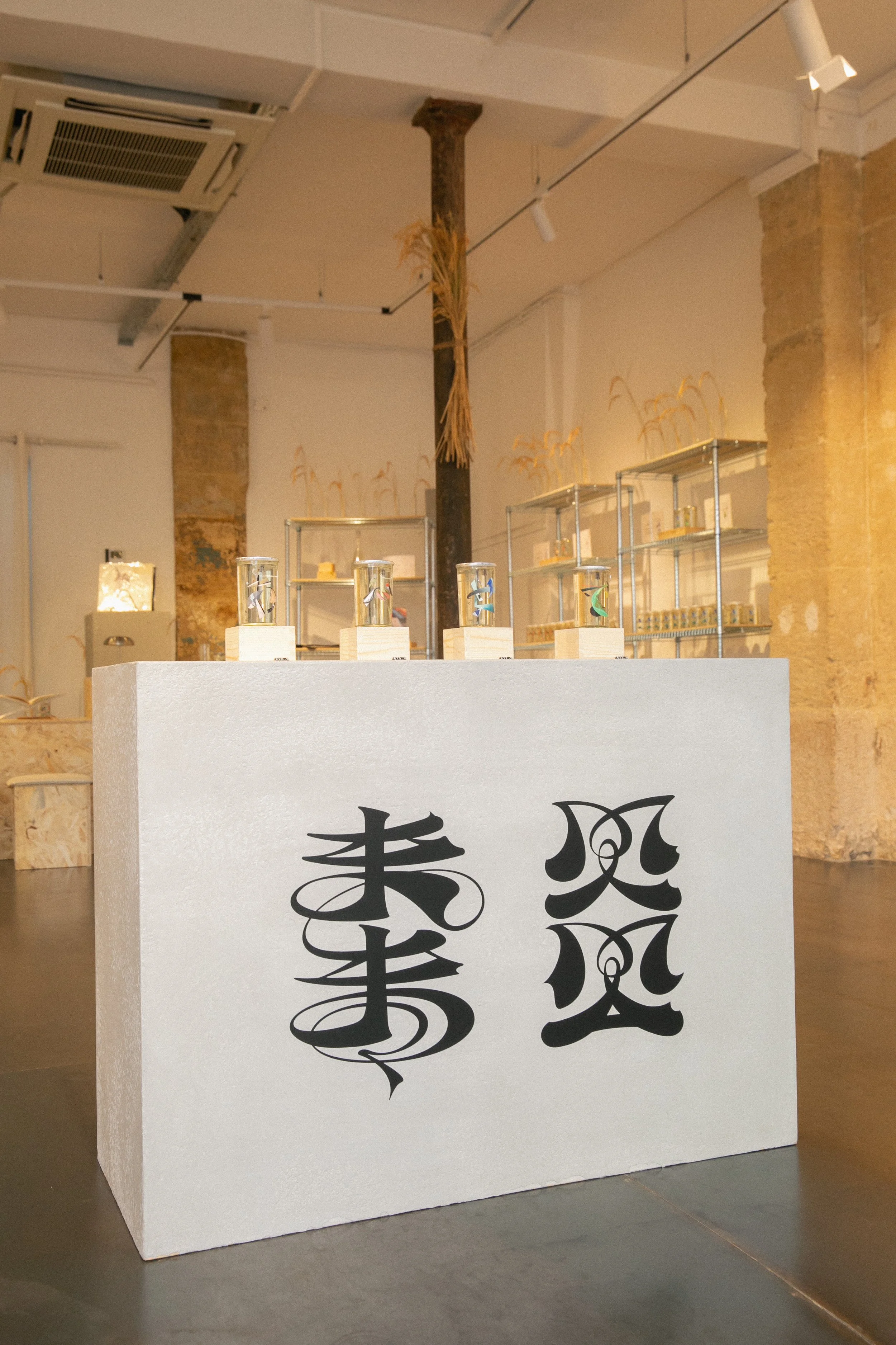 A white display stand with black Asian calligraphy characters in the foreground inside a room with industrial-style exposed pipes and a brick wall column in the background.