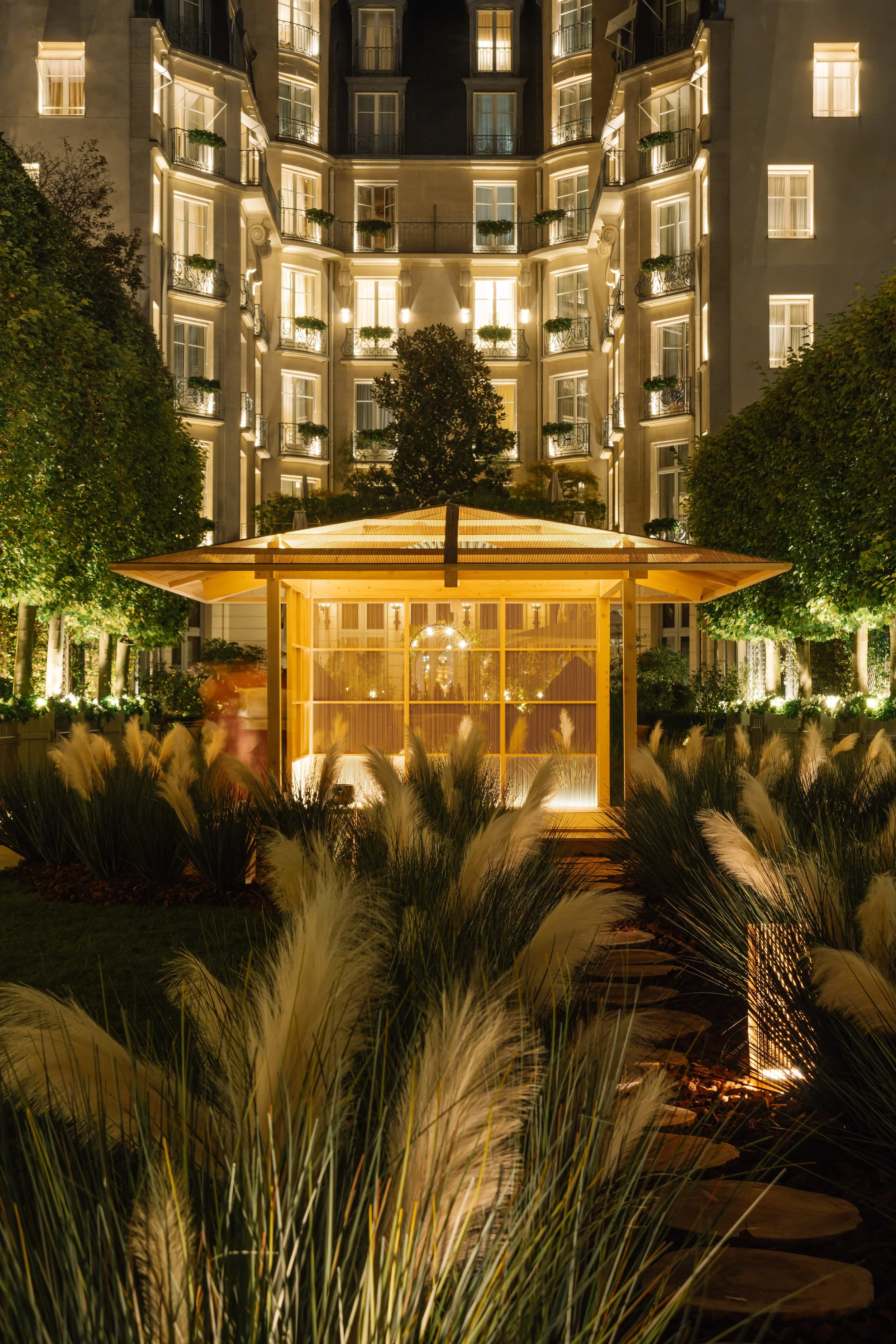 Night shot of a multi-story hotel building with lit windows and balconies, seen behind a landscaped garden with tall, illuminated grasses and trees, and a wooden structure in the foreground.
