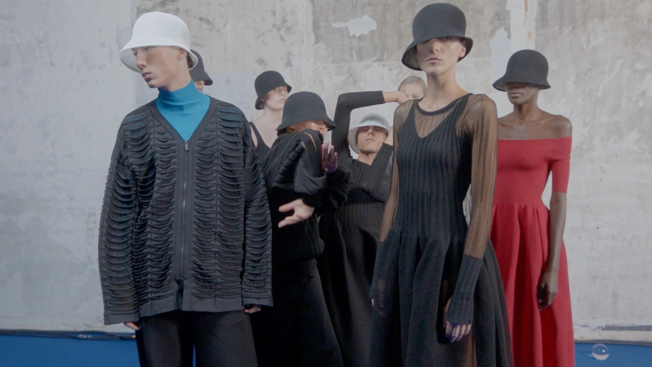 Group of five women standing against a concrete wall, wearing stylish clothing and hats, posing in a fashion photoshoot.