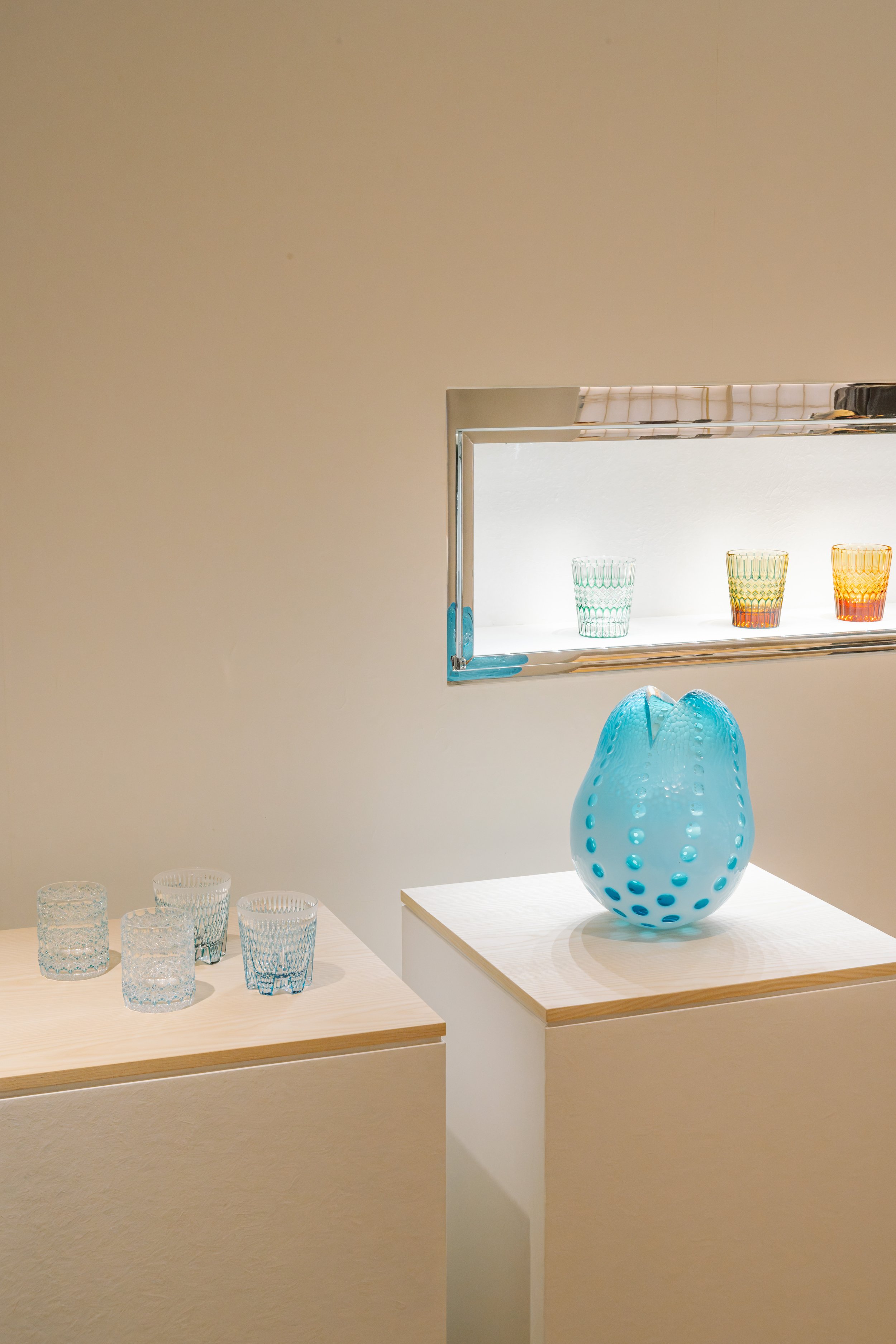 Display of colorful glassware, including a large blue vase with holes, and several smaller glasses in various colors and patterns, arranged on white pedestals and tables in a gallery setting.