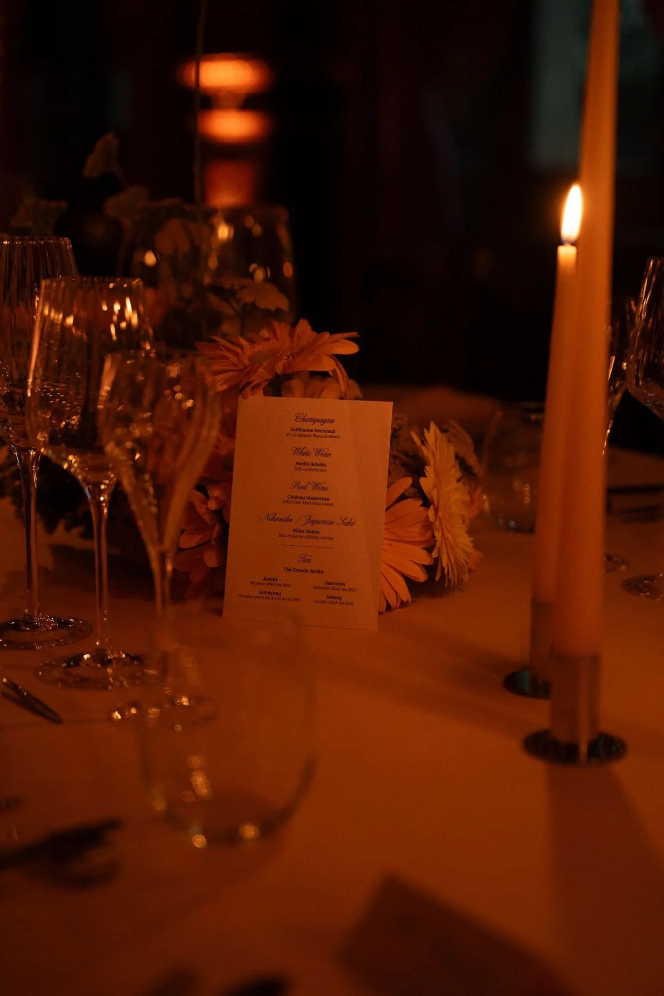 A dimly lit dining table with wine glasses, a candle, flowers, and a menu card in a fancy setting.