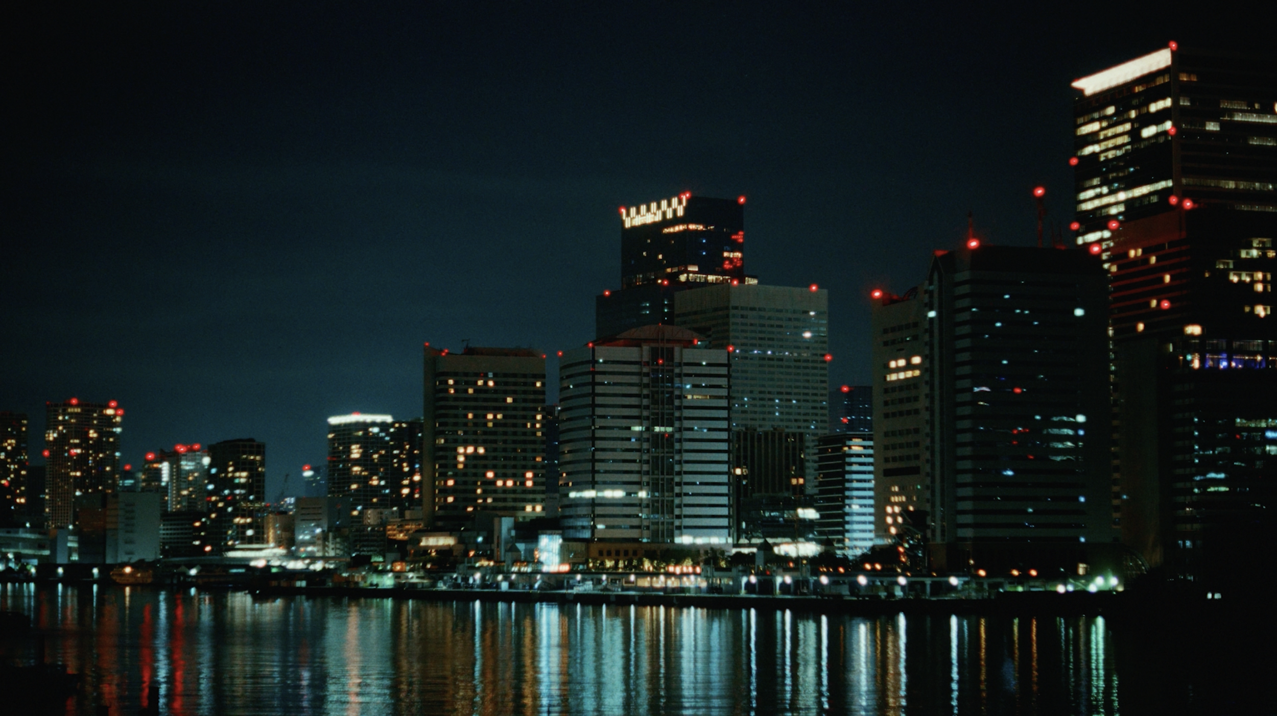Nighttime city skyline with tall illuminated buildings reflecting on the water.