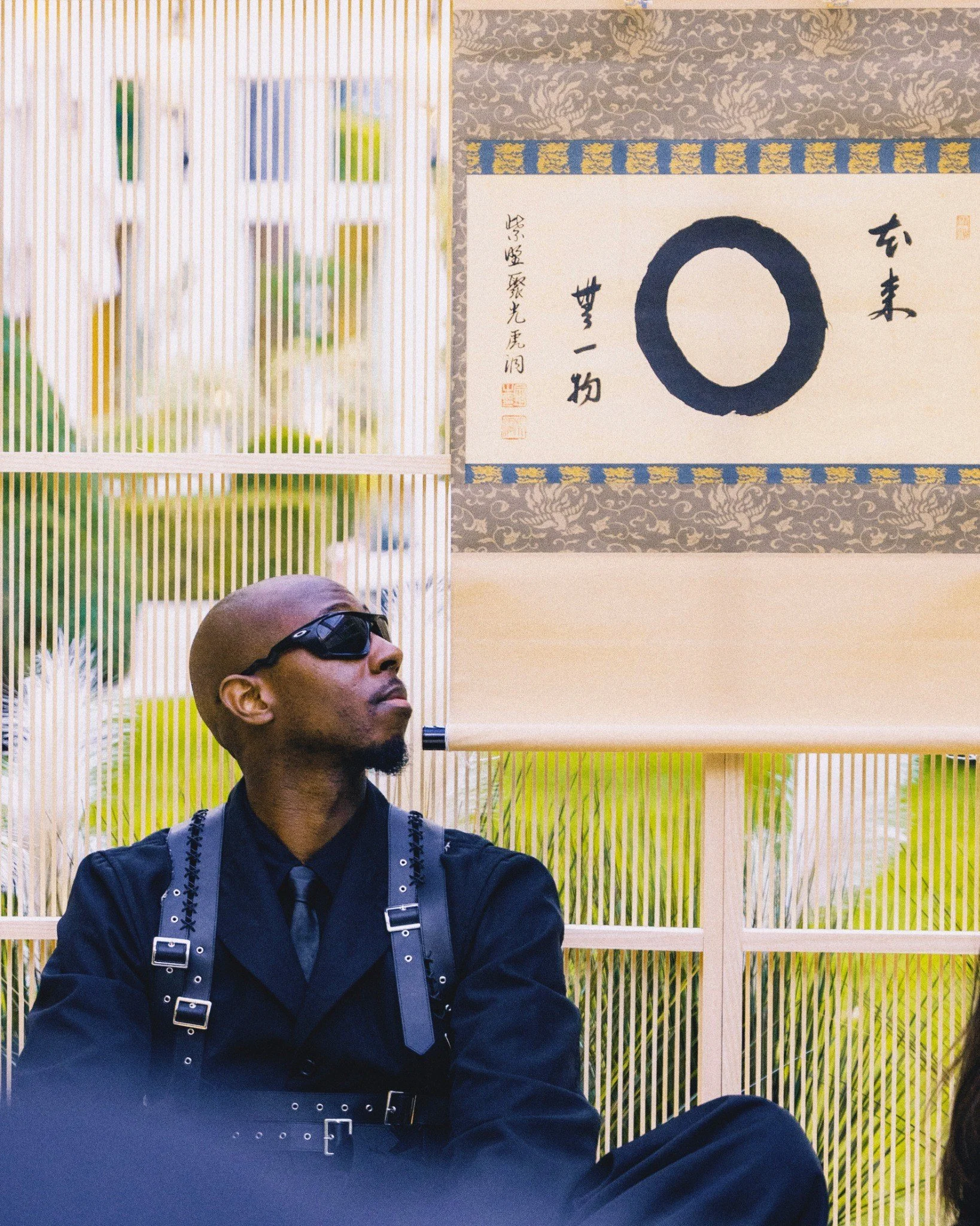 Bald man wearing black sunglasses, black shirt, and gray suspenders with silver buckles, sitting outdoors with yellow-green plants and bamboo screen behind him