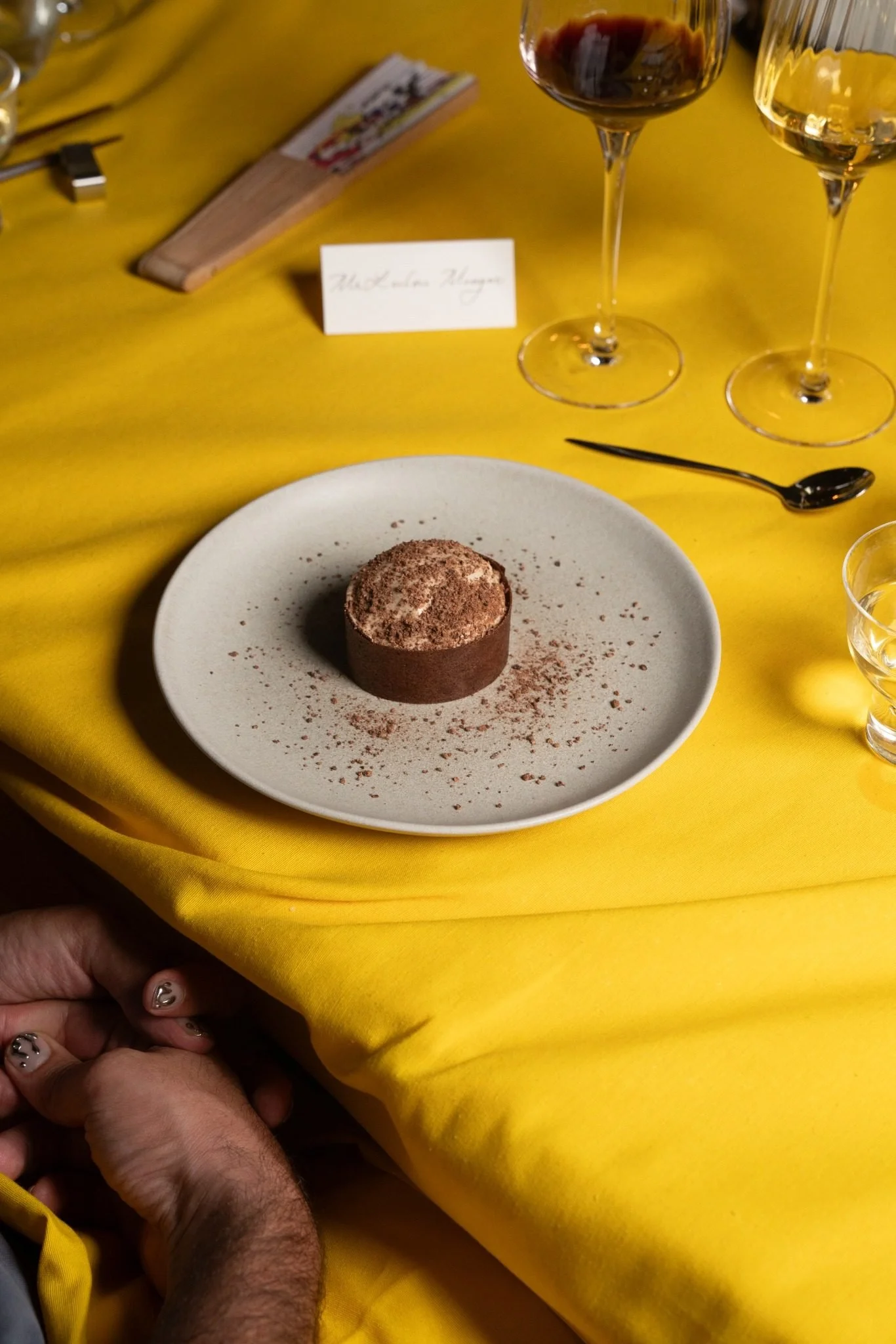 A plate with a chocolate dessert covered in cocoa powder on a yellow tablecloth, alongside two glasses of red and white wine, a name card, and a hand holding a napkin.