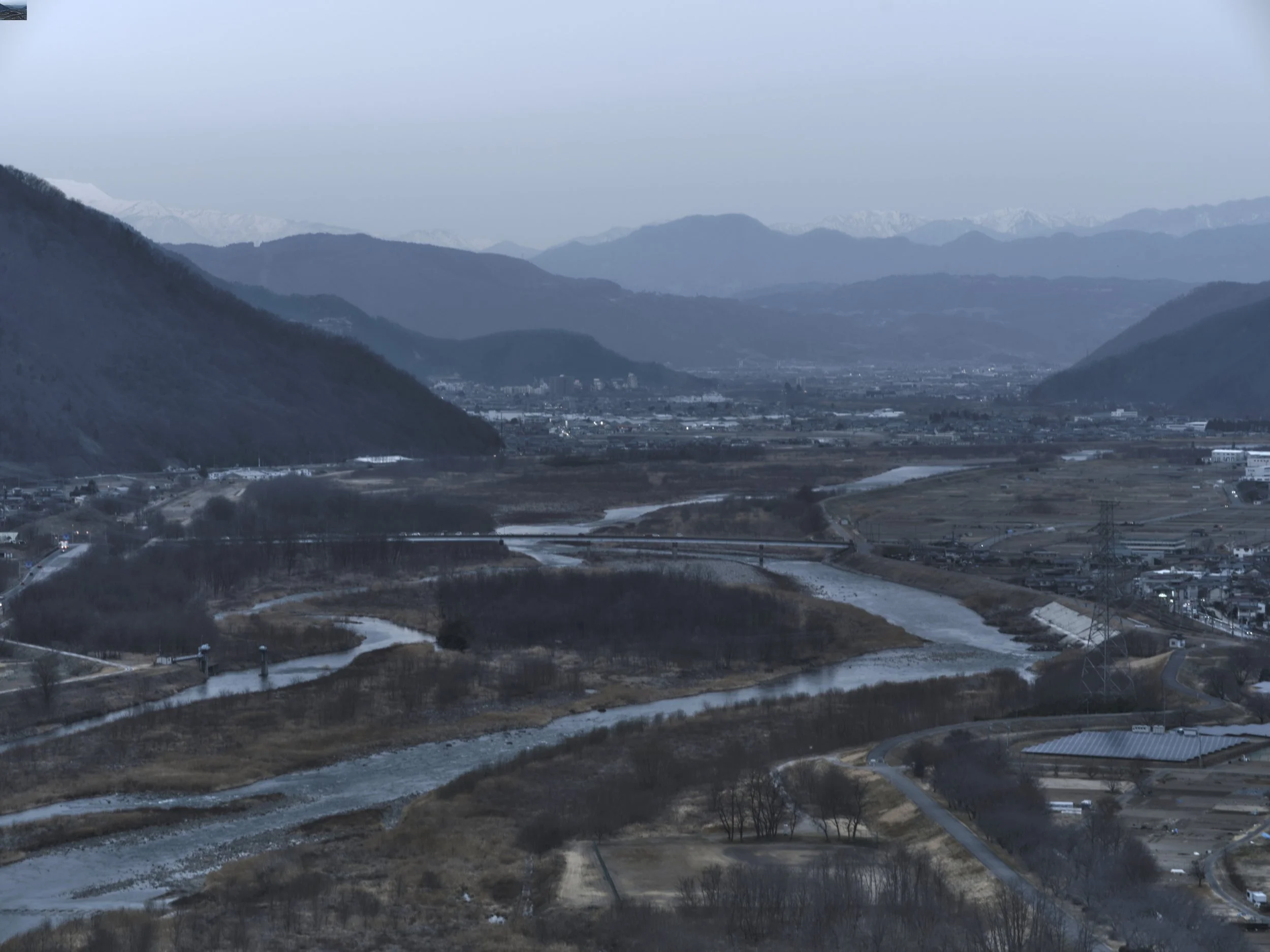 A mountainous landscape with snow-capped peaks in the background, rolling hills in the middle, and a rural area with roads and small buildings in the foreground.