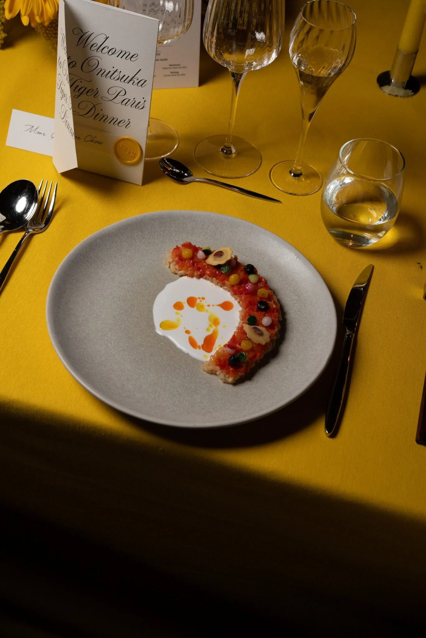 A plate with a crescent-shaped dish of rice topped with diced tomatoes, herbs, a dollop of yogurt or sour cream, and garnished with small colorful candies or pearls. The table has a yellow tablecloth, and there are empty wine glasses, a glass of wate