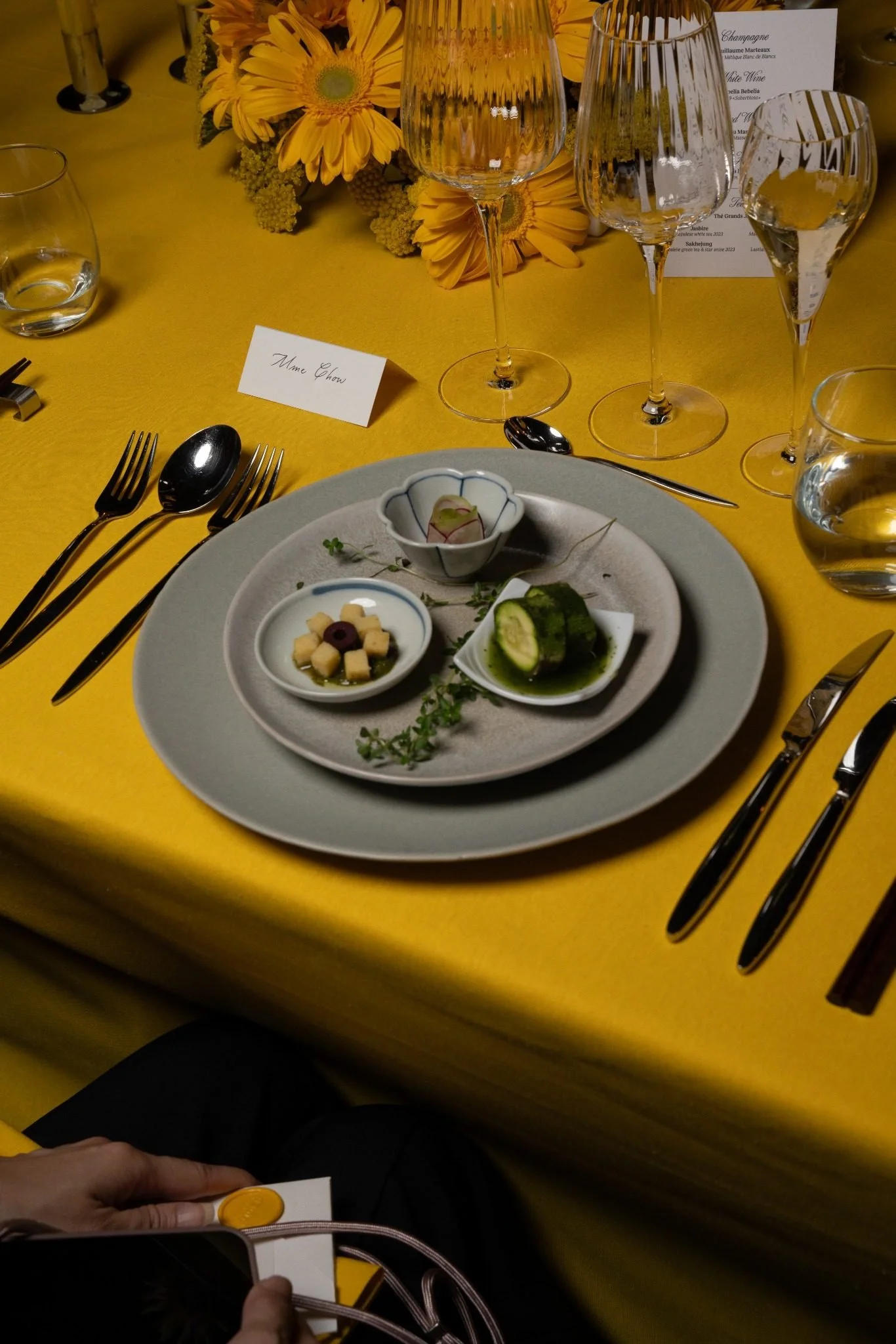 A table set for a formal dining event with a yellow tablecloth, wine glasses, cutlery, a small plate with appetizer, and a floral centerpiece with yellow flowers. There are also place cards, one of which reads 'Mme Chow,' and a menu card in the backg