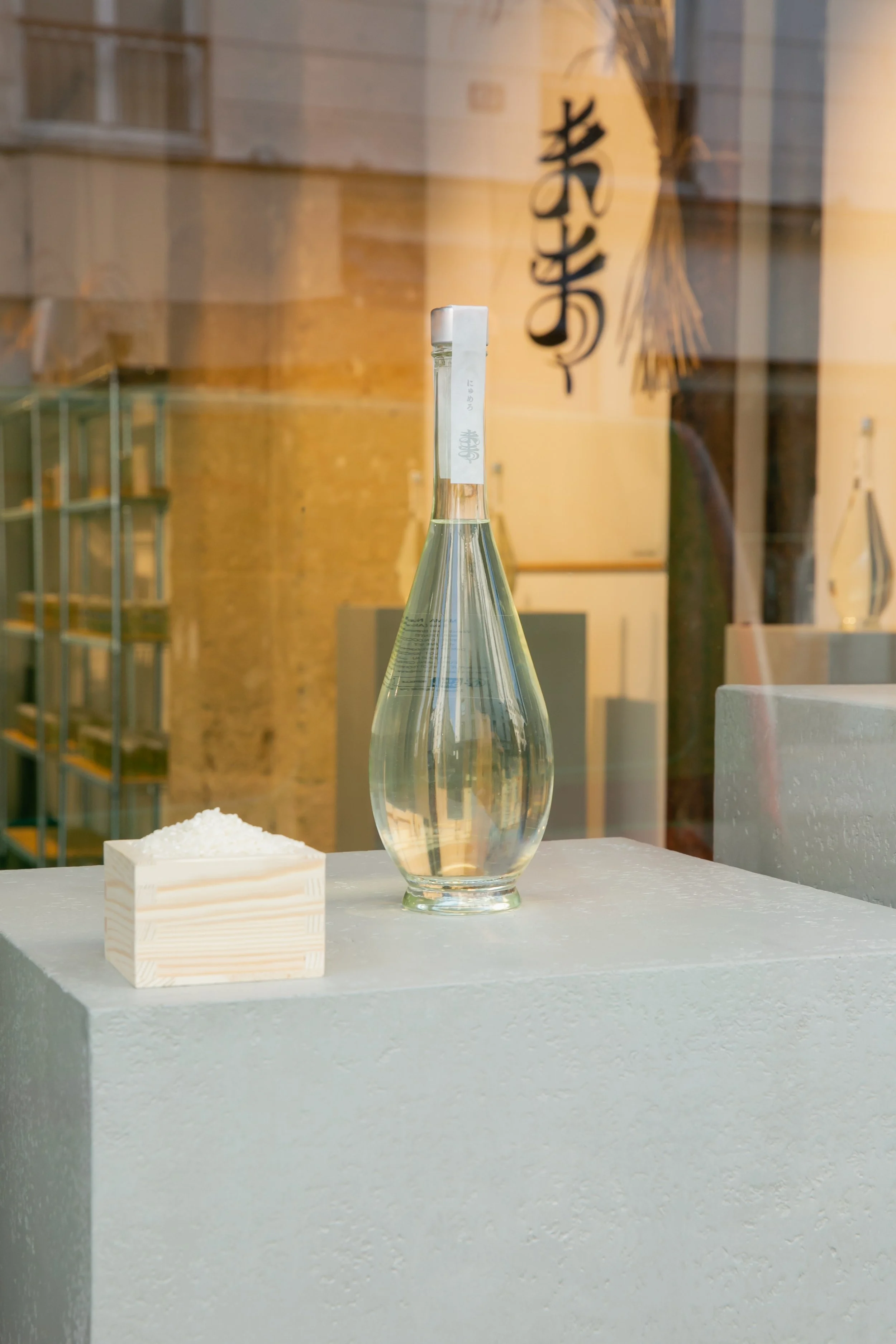 A tall clear glass bottle filled with a transparent liquid, placed on a white textured surface. Next to it is a small wooden container with white grains inside. The background features a window with reflections of a Japanese paper lantern with Japane