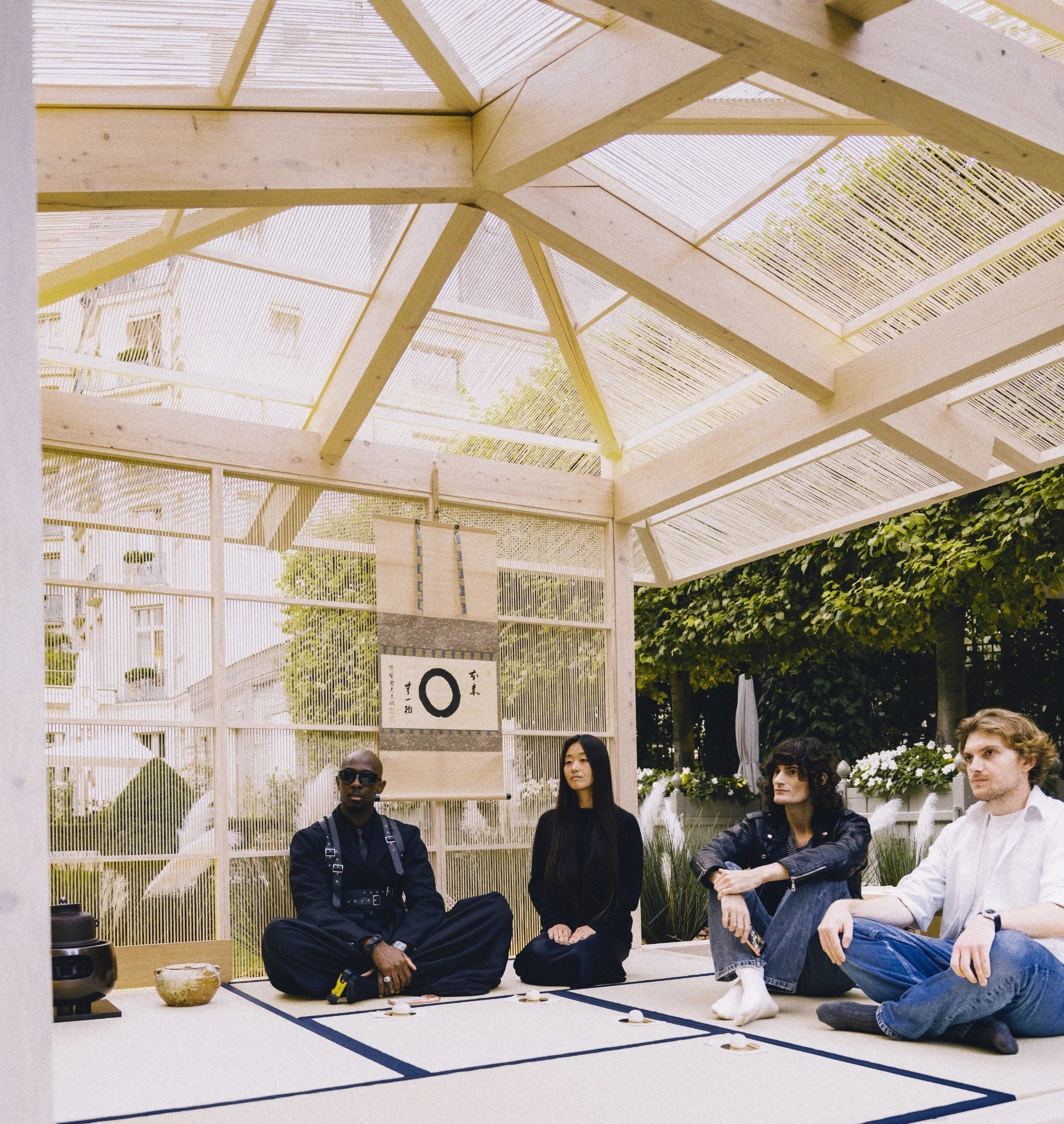 People sitting on a tatami mat in a traditional Japanese room with a wooden frame ceiling, participating in a tea ceremony, with some wearing glasses and casual clothing, and a decorative hanging scroll in the background.