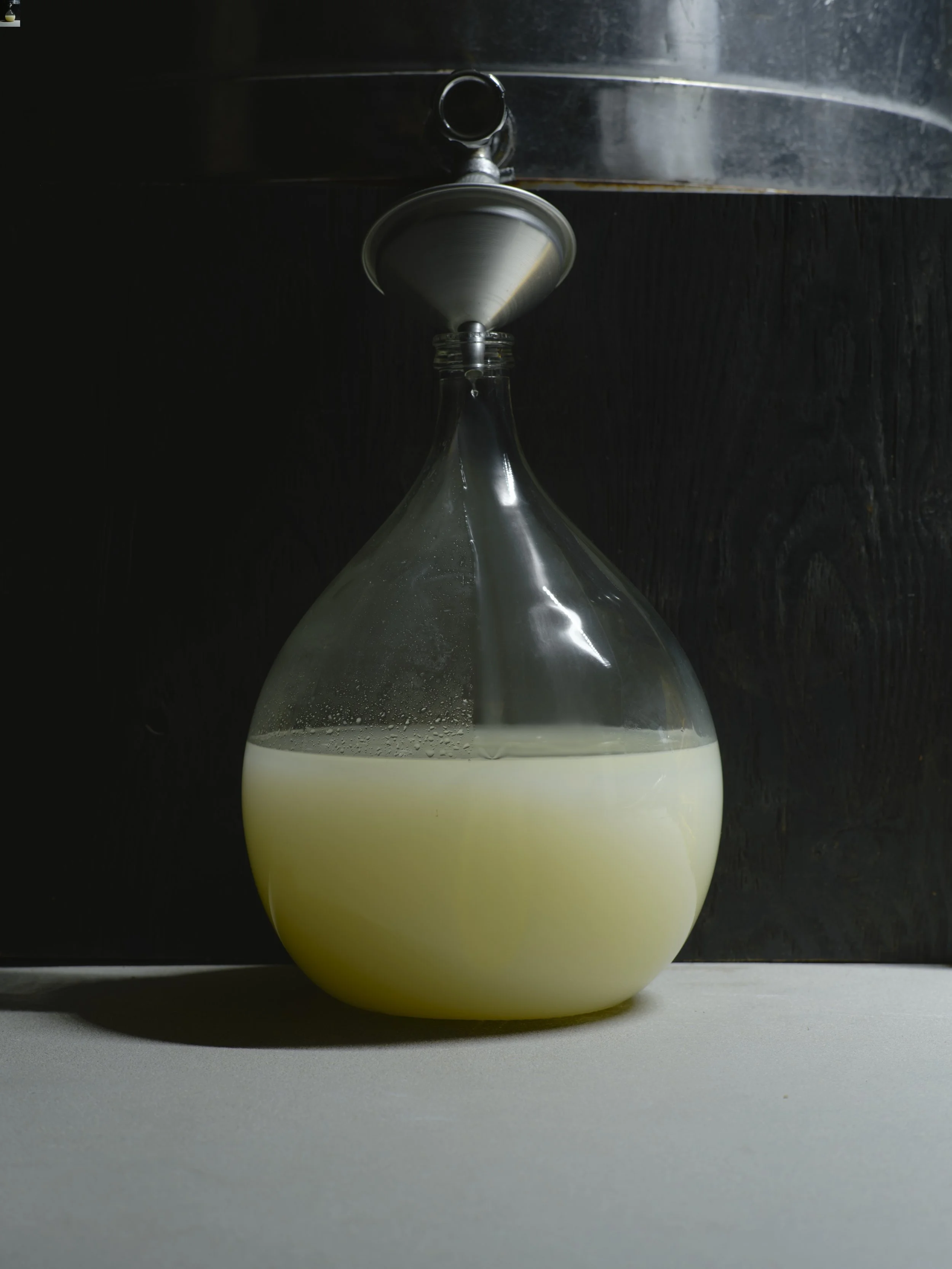 A glass bottle with a metal lid, pouring a white liquid into a container on a dark surface, with a dark textured background.