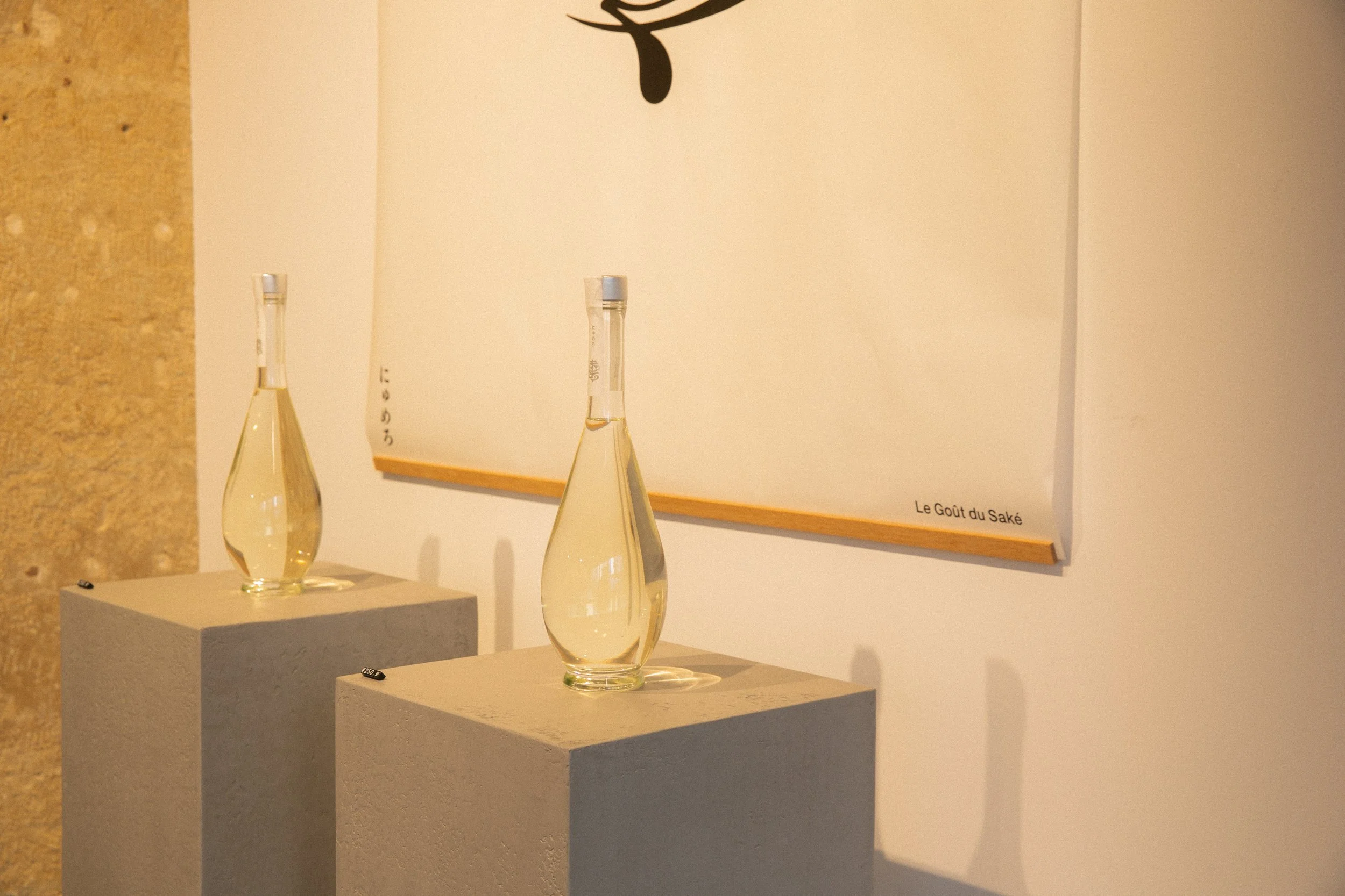 Three glass bottles of sake on stone pedestals in an art gallery, with a white wall featuring a black abstract design and the words "Le Goût du Saké".
