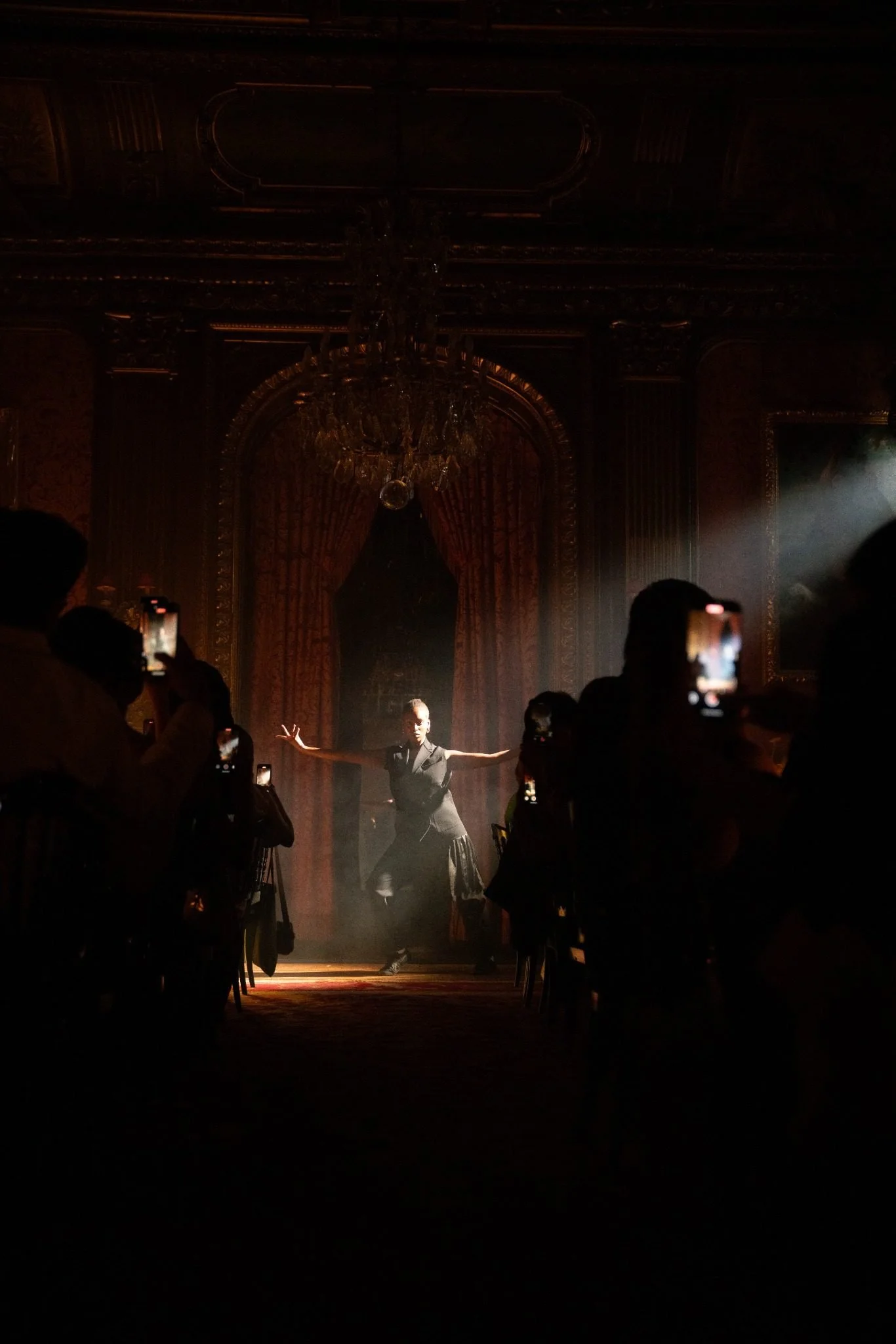 A performer with arms extended standing on a stage in a dimly lit, ornate room with a chandelier and heavy curtains, while audience members record the performance with their phones.